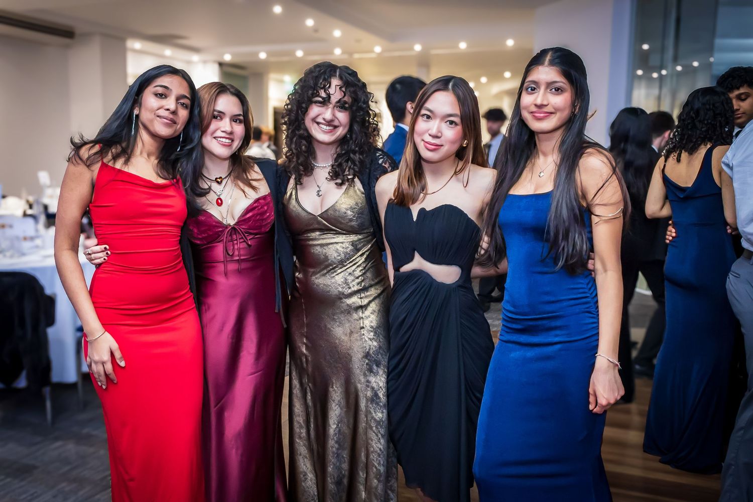 Five women in elegant formal gowns posing together at a school ball.