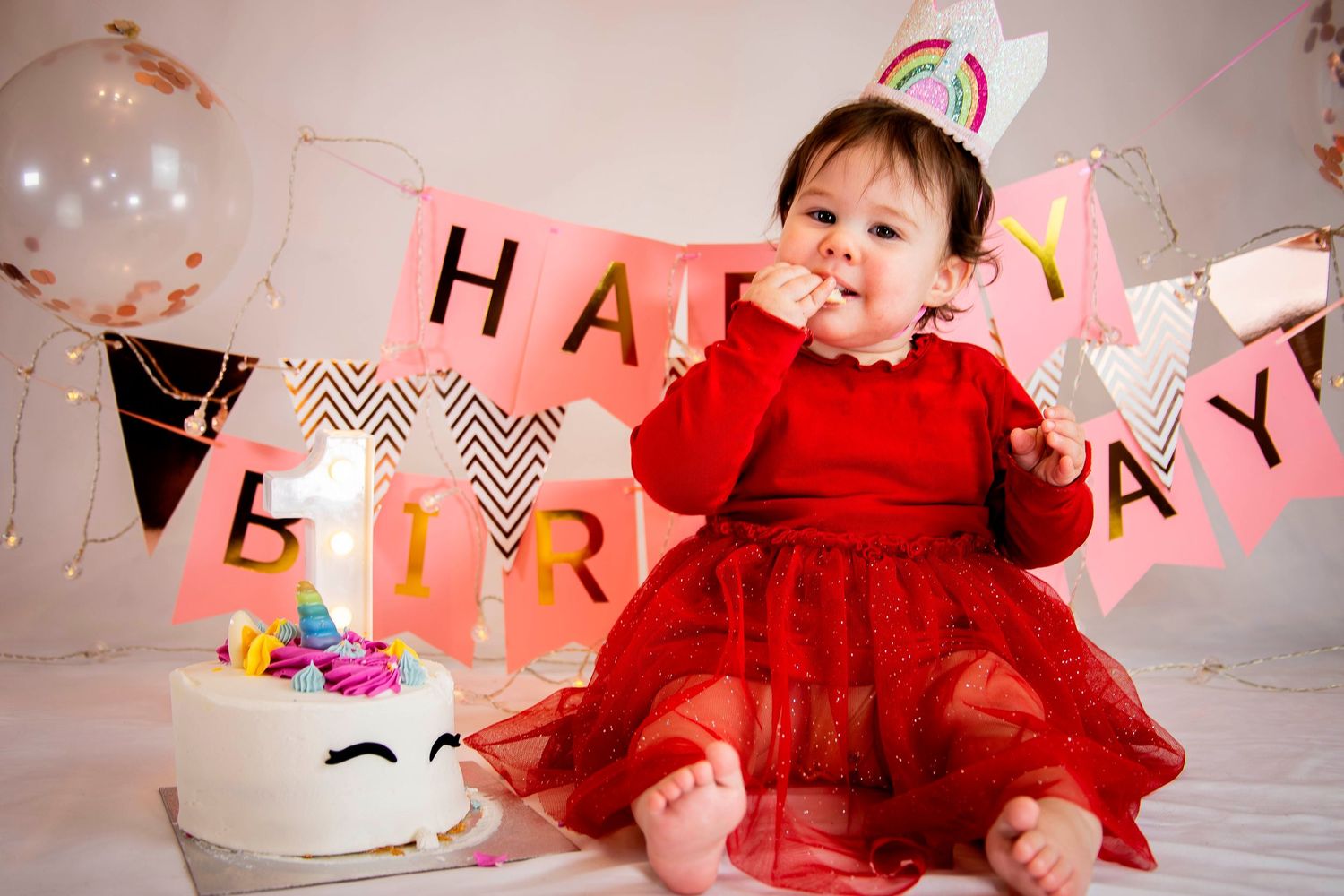 Adorable baby girl in red dress sitting before Happy Birthday banner with unicorn cake on her first birthday.