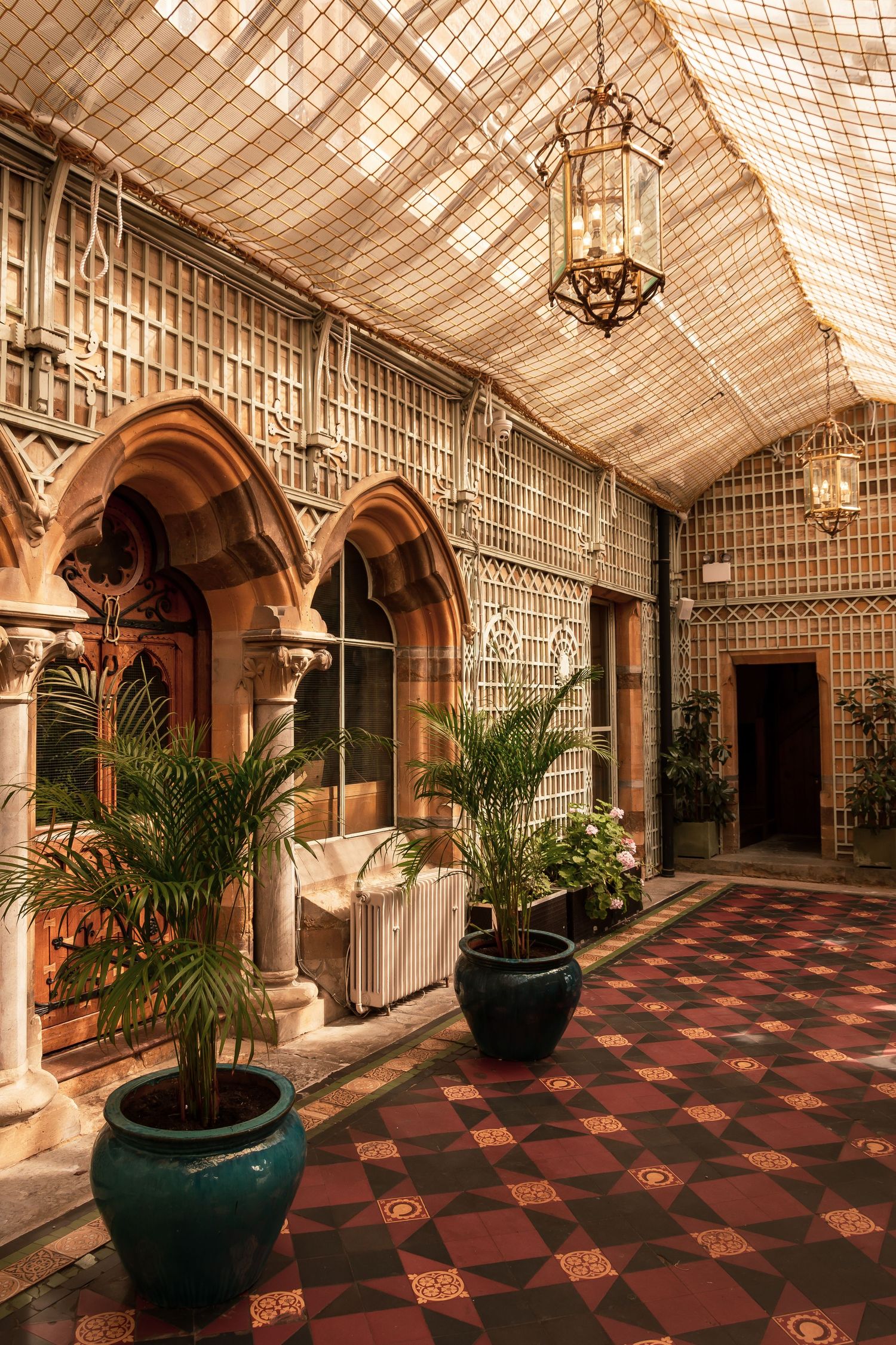 Ornate Victorian conservatory corridor with Gothic arches, tropical potted palms, and decorative tiled flooring.