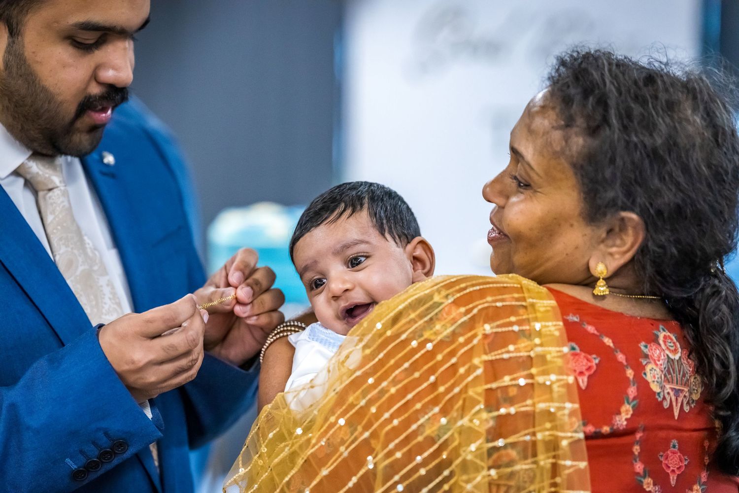 Smiling baby in white christening outfit held by elderly woman while man in blue suit reaches out.
