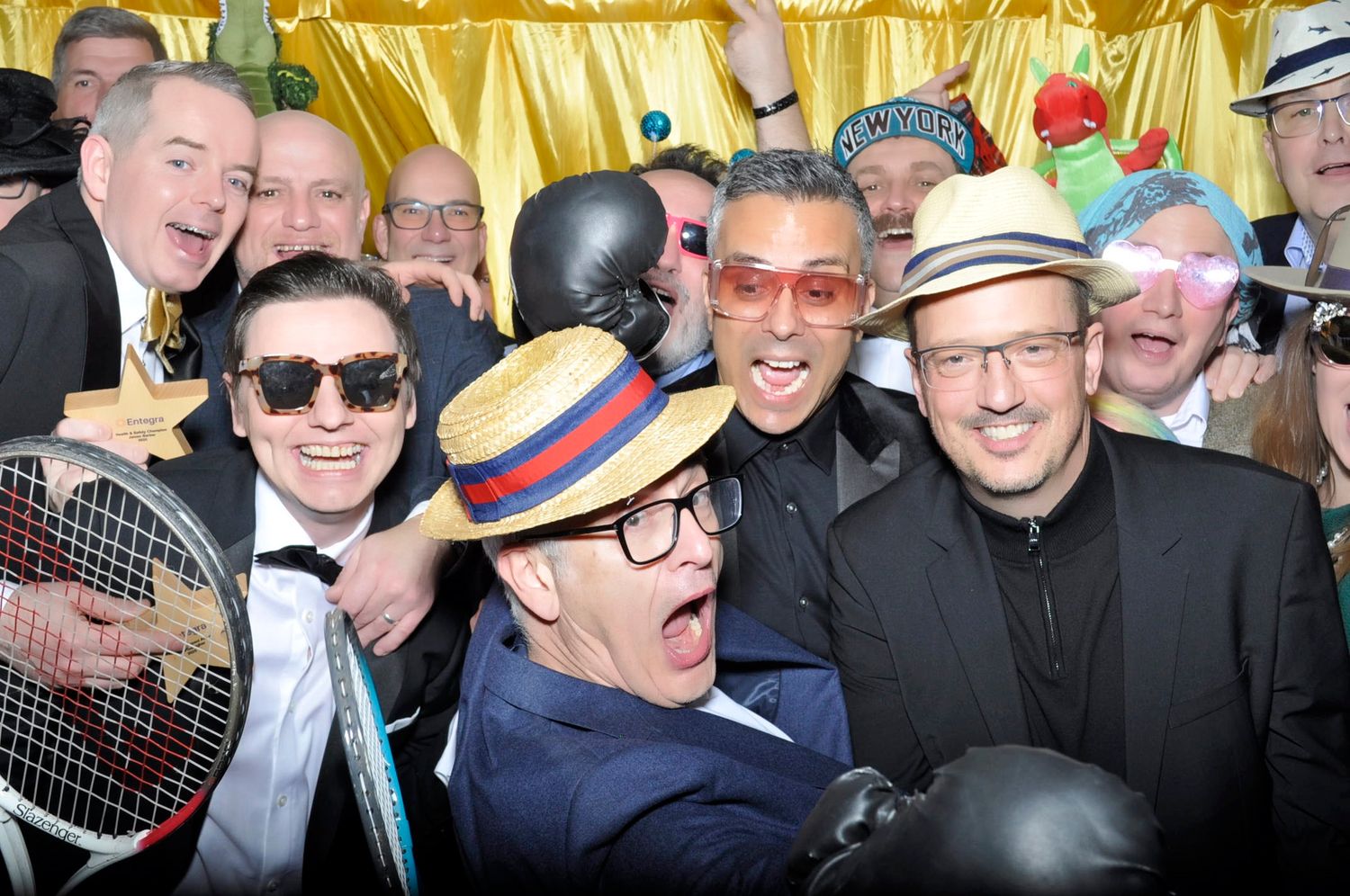 A group of men in hats and sunglasses pose playfully with props at a festive party photo booth.