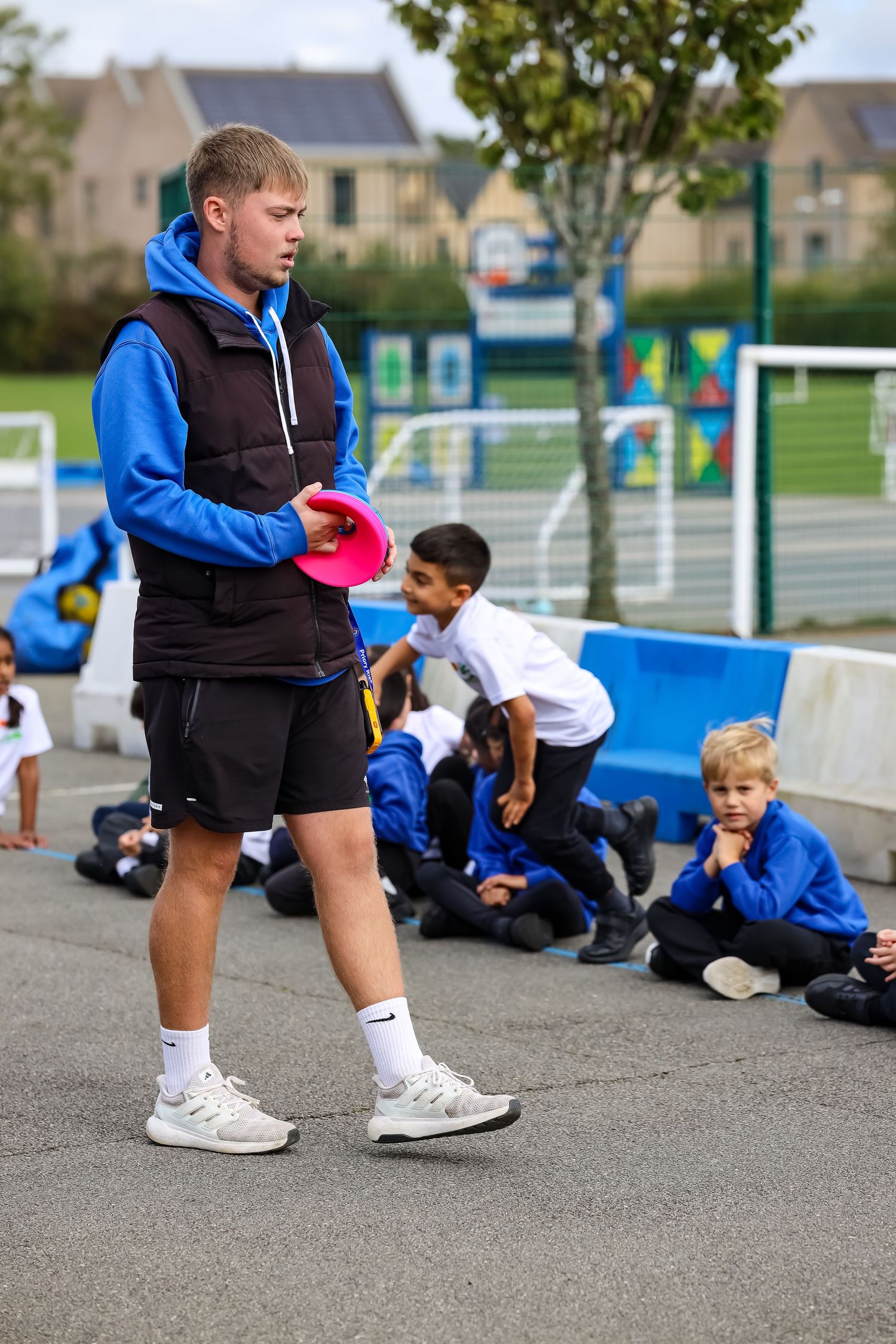 PE teacher holding a pink disc while students sit on a playground during sports lesson.