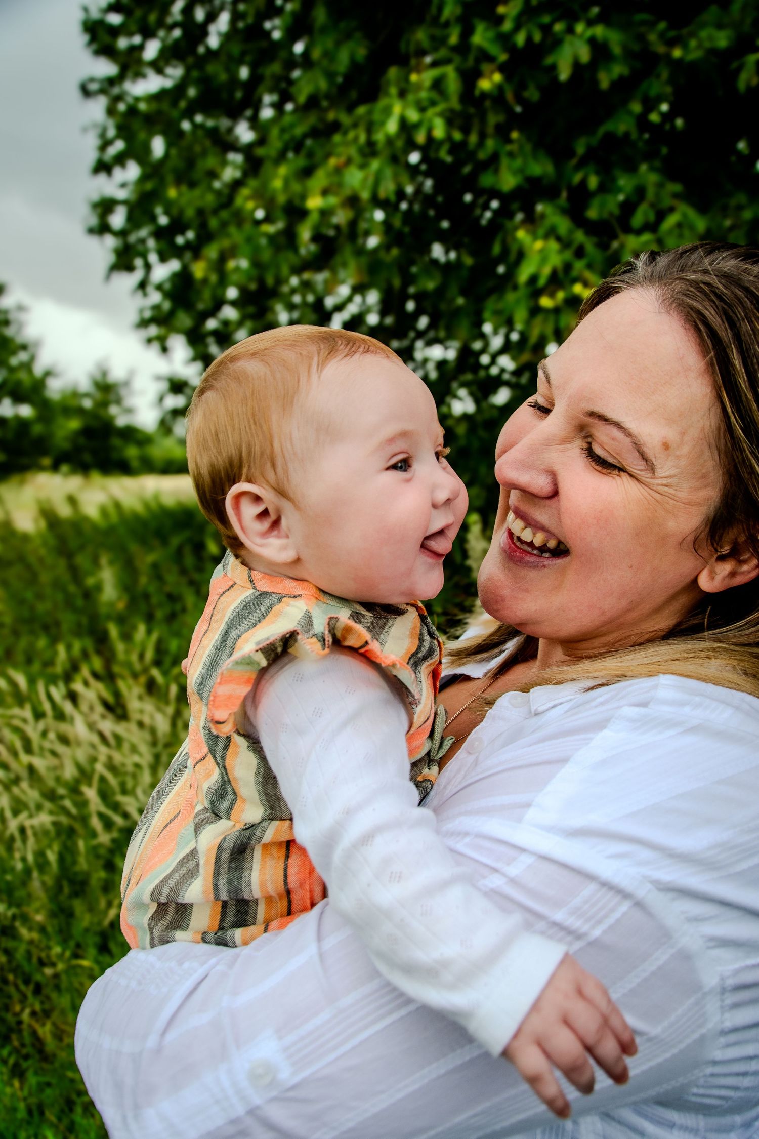 Laughing mother holding a giggling baby outdoors near green trees in a joyful moment.