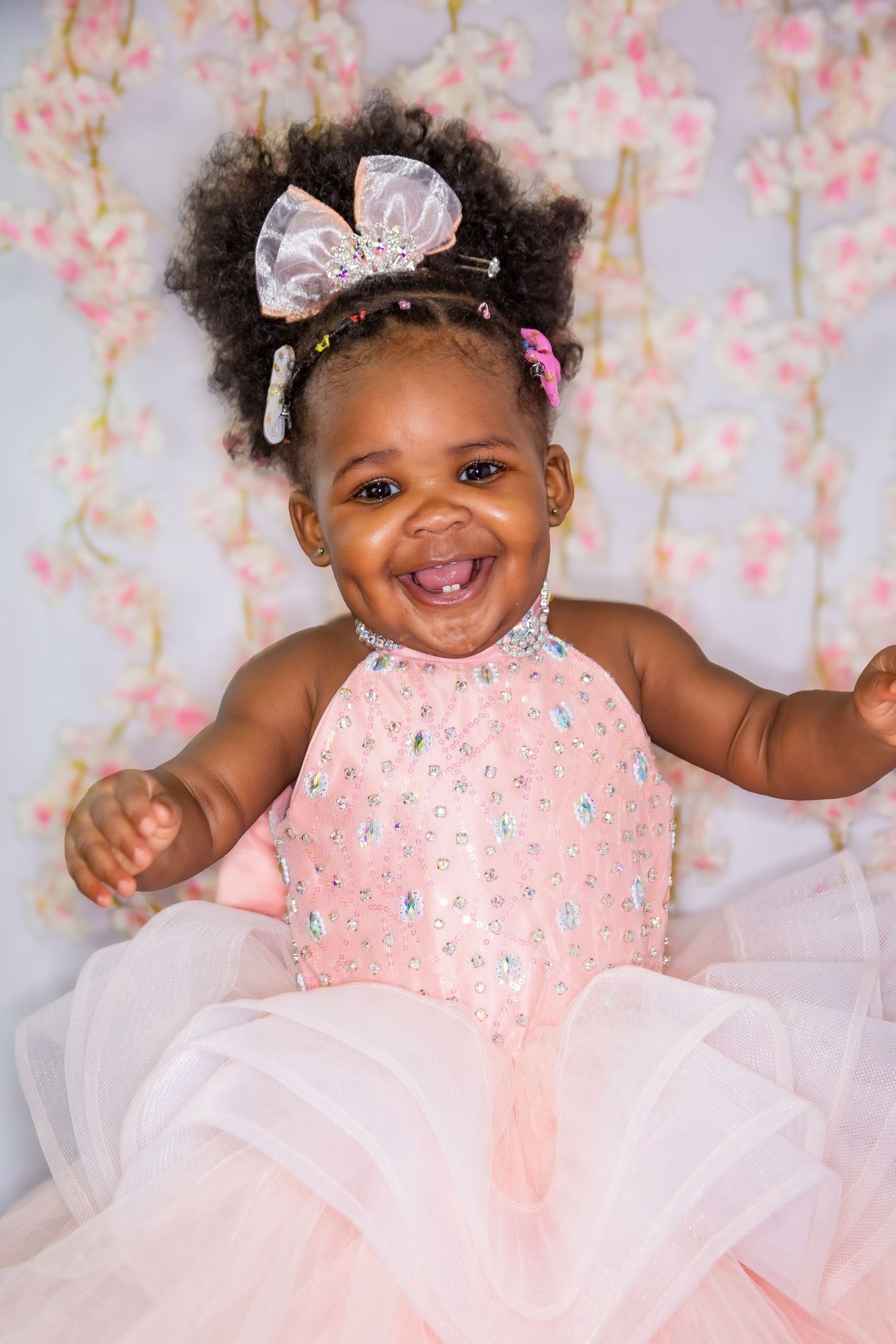 Joyful baby girl in pink rhinestone dress with bow hair accessories laughing against floral backdrop.