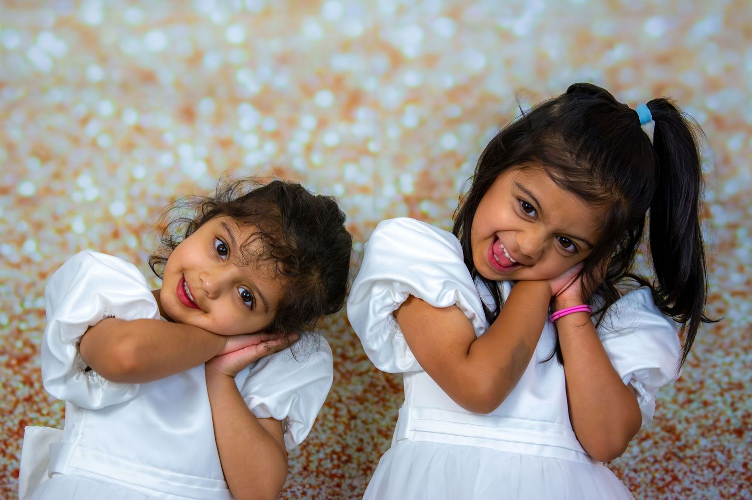 Two young sisters in matching white dresses posing playfully against a bokeh background.