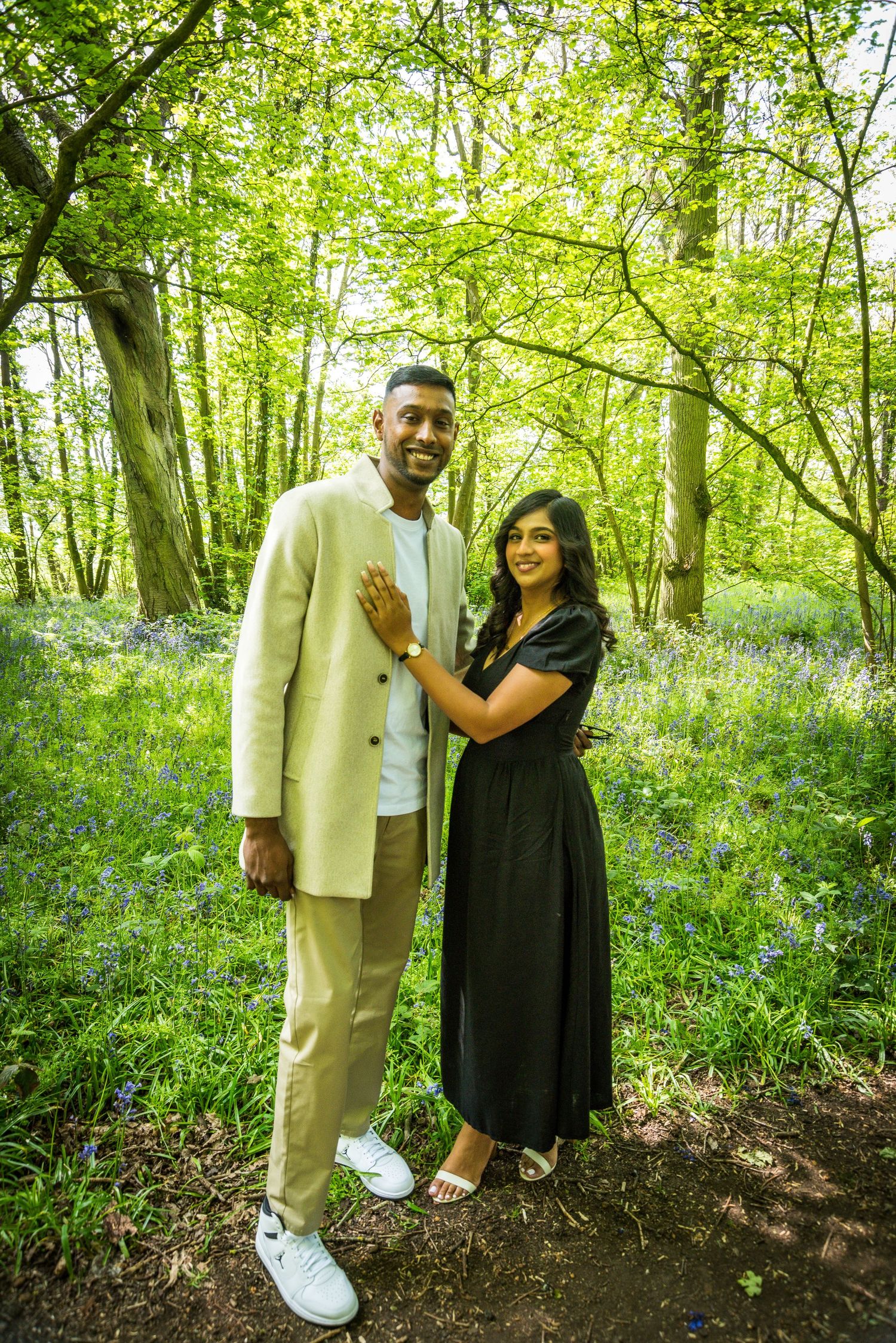Couple posing together in a bluebell woodland, man in beige suit and woman in black dress smiling.
