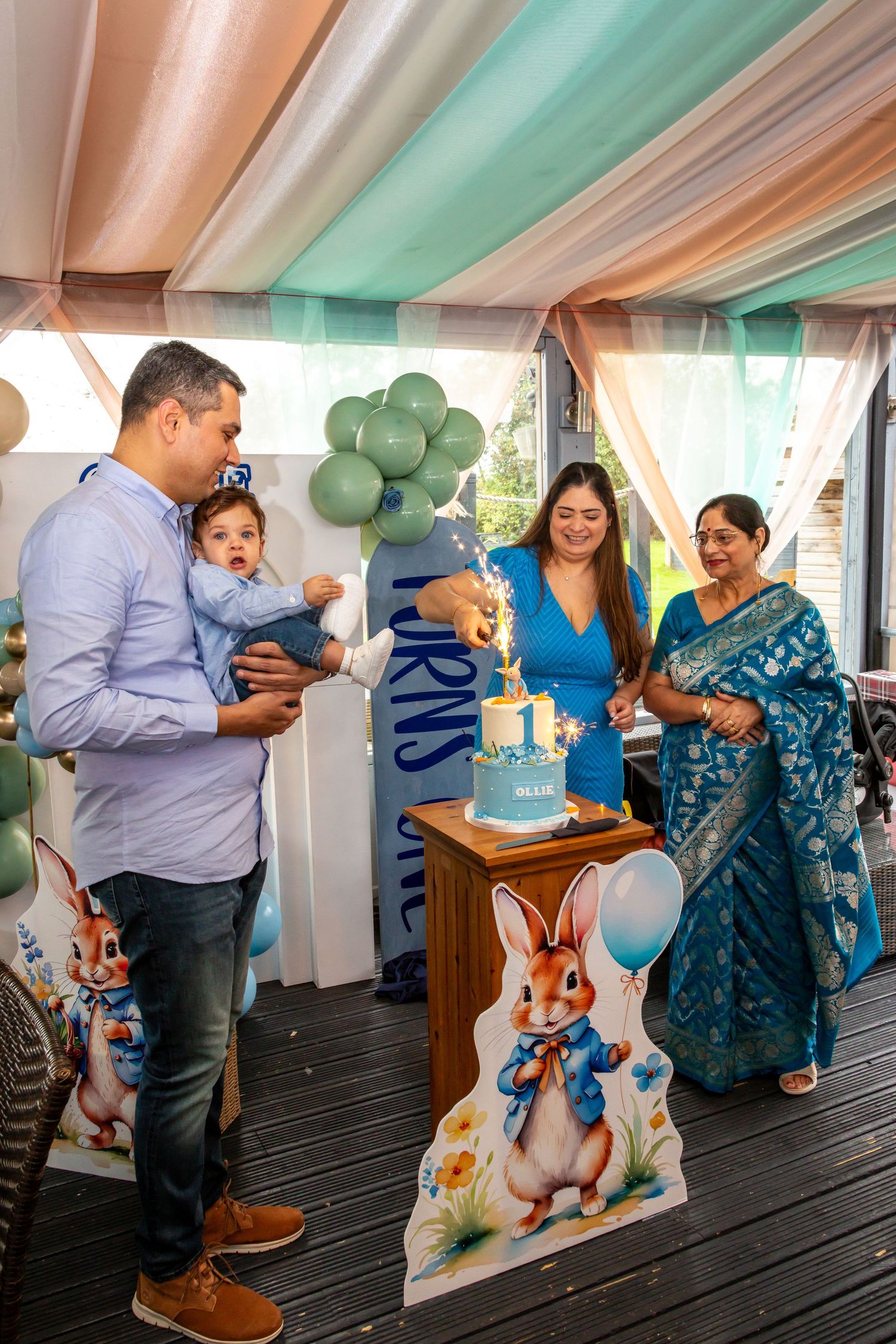 Family celebrating baby Ollies first birthday with a blue Peter Rabbit cake and sparkler.