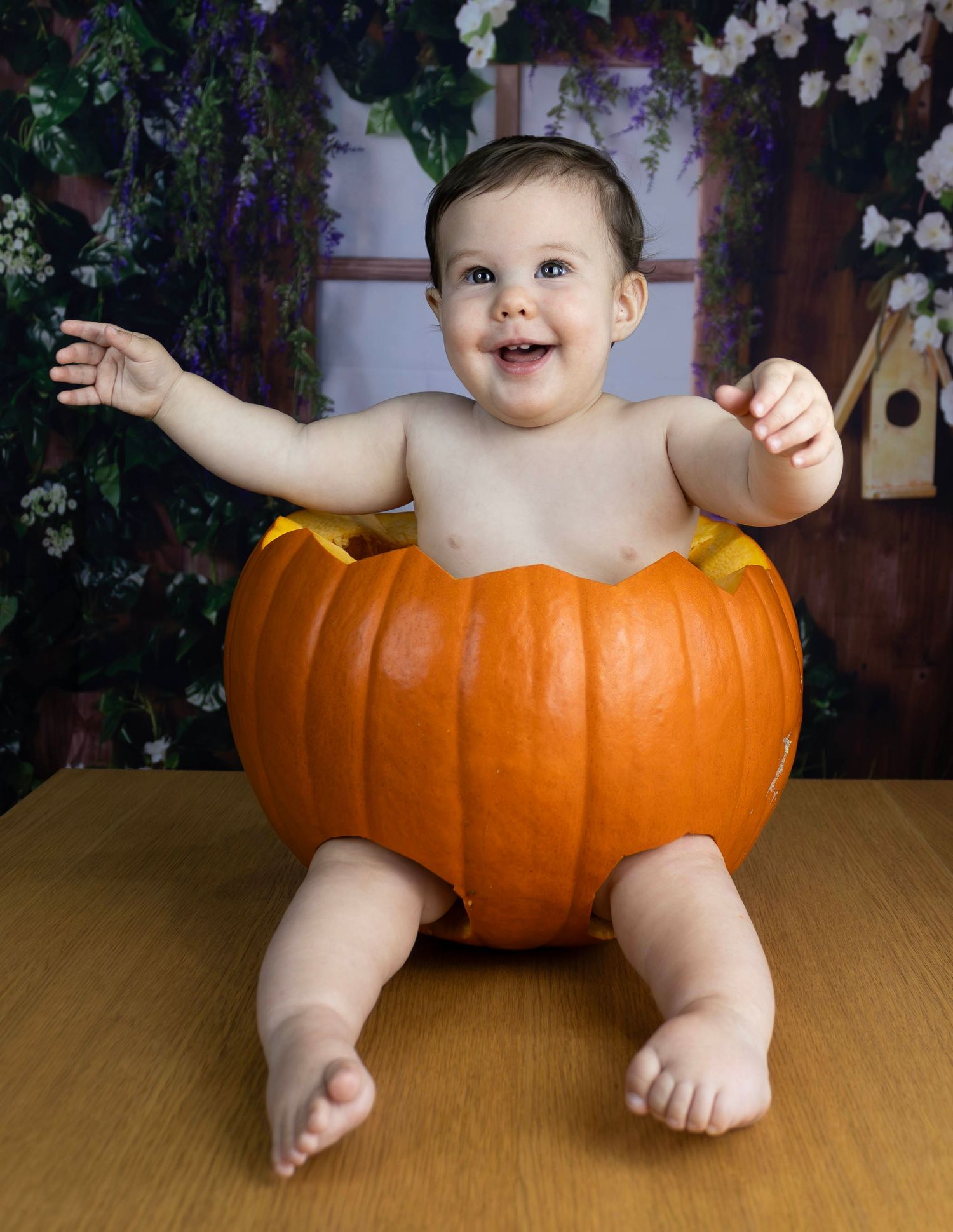 Adorable smiling baby sitting inside a carved Halloween pumpkin with floral backdrop.