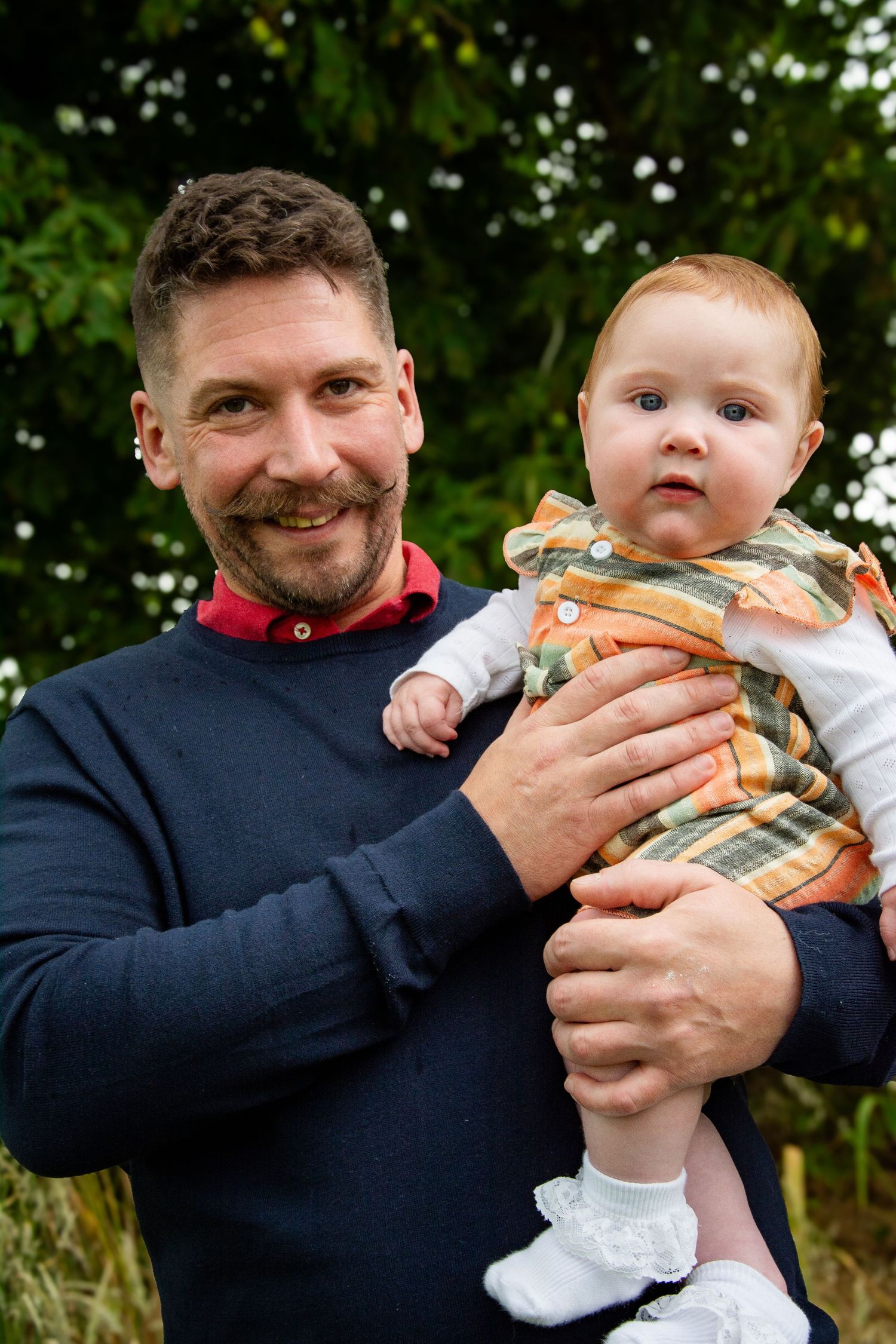 Bearded man in navy sweater smiling while holding an alert baby in a plaid outfit outdoors.