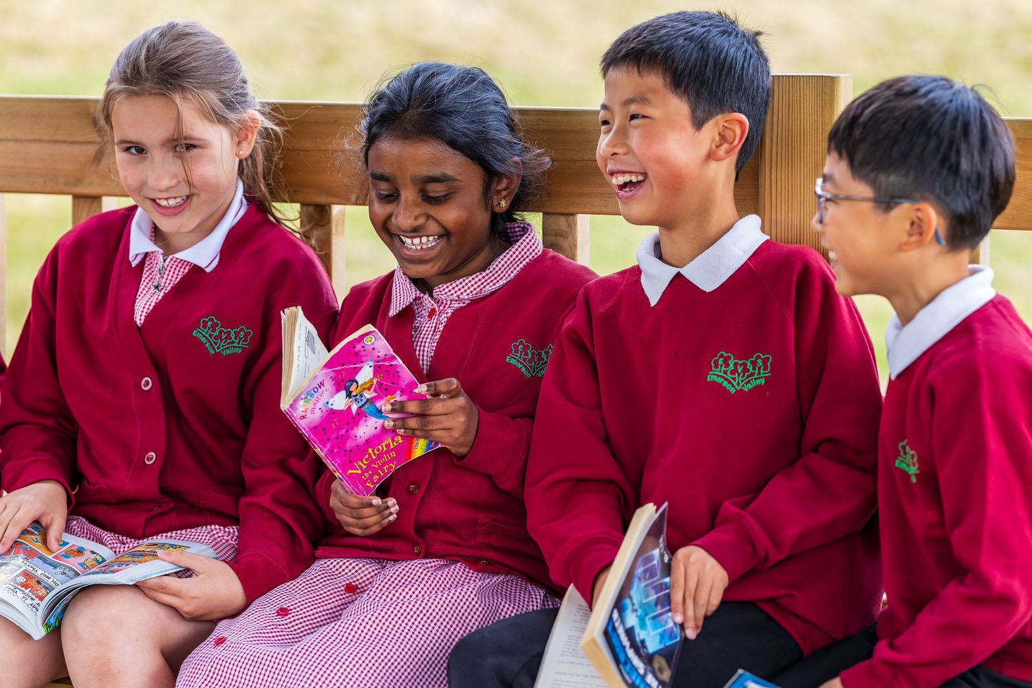 Four happy primary school children in red uniforms reading books on a bench.