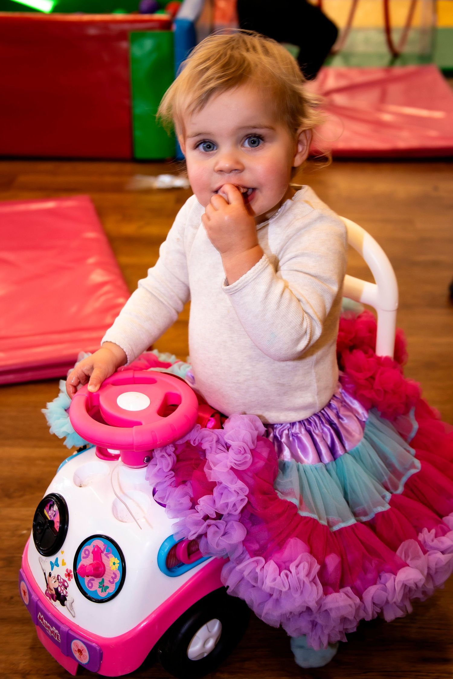 A cute toddler girl in a pink tutu skirt plays with a Minnie Mouse ride-on toy car.