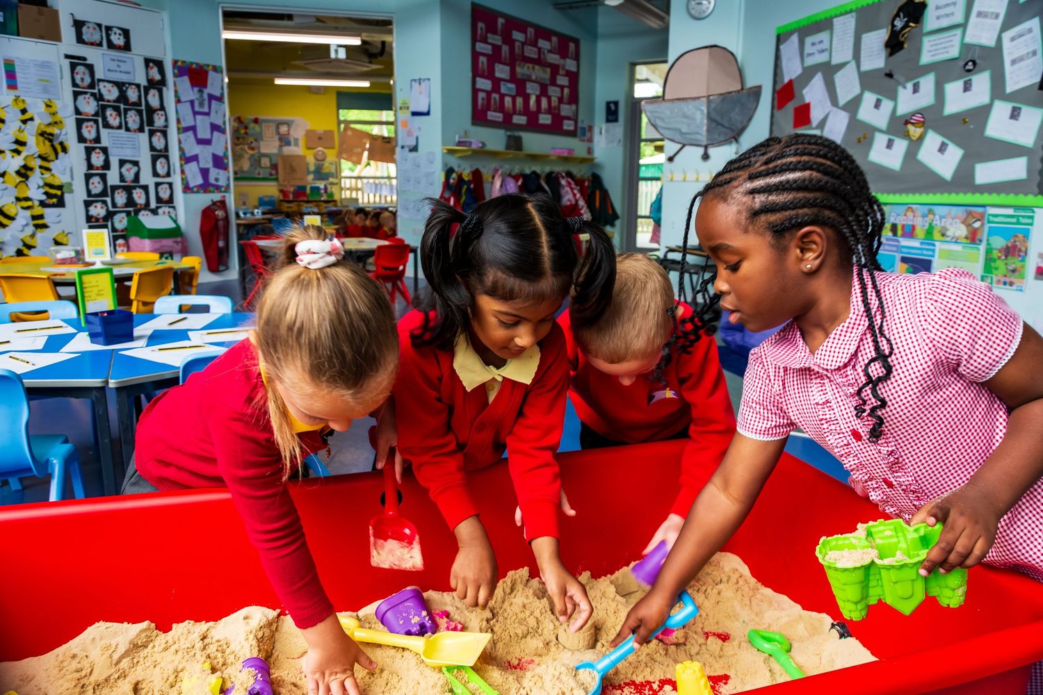 Four young children in red uniforms playing together in a sandbox at an elementary school classroom.
