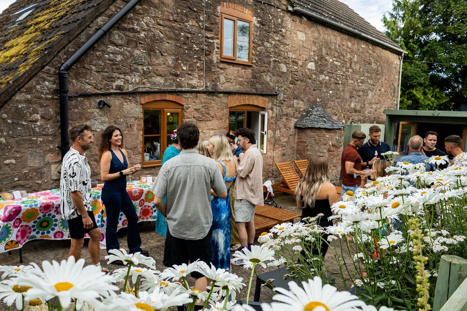 Group of people socializing outside a rustic stone cottage surrounded by white daisy flowers.