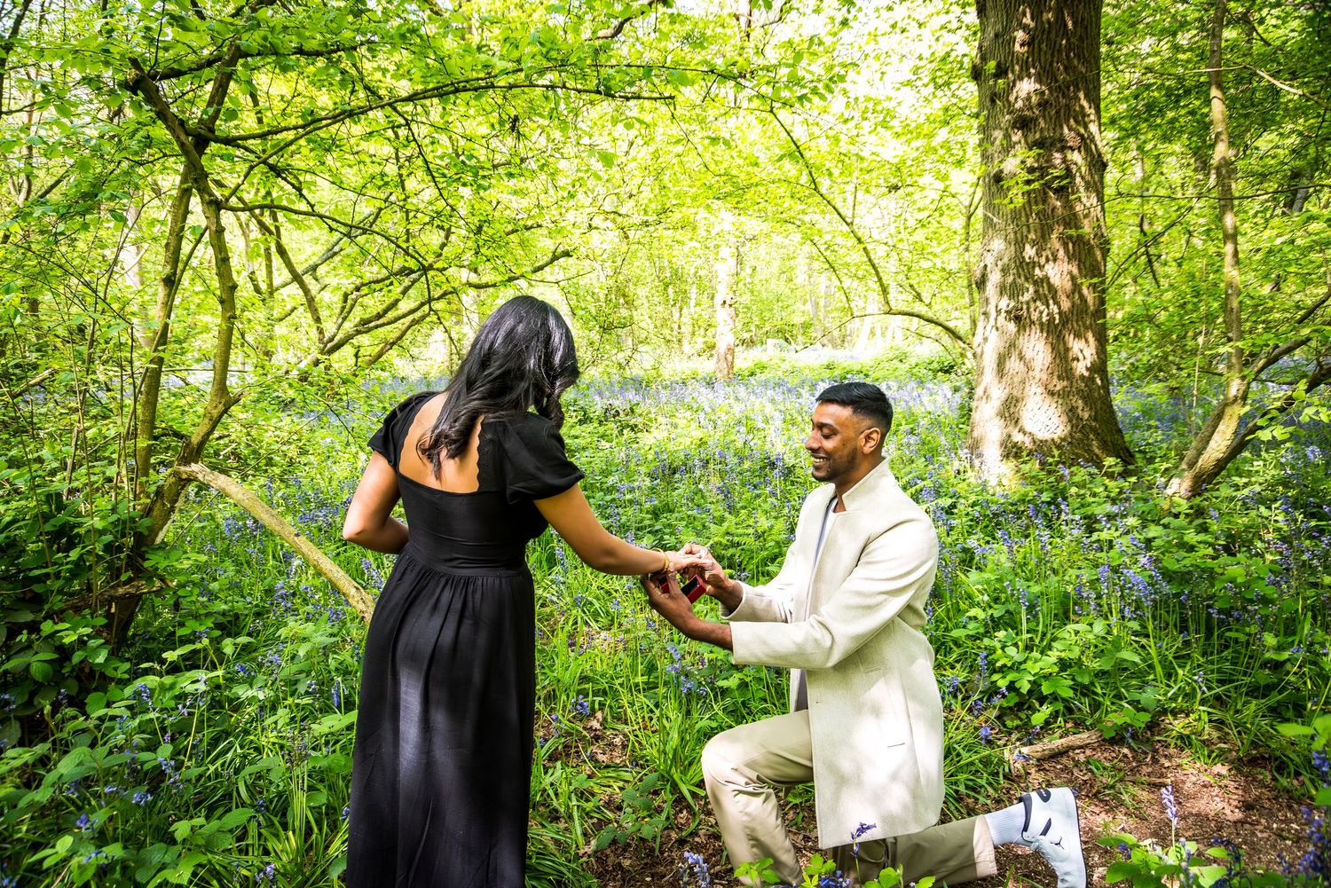 Man kneeling on one knee proposing to woman in black dress in a lush green woodland with bluebells.