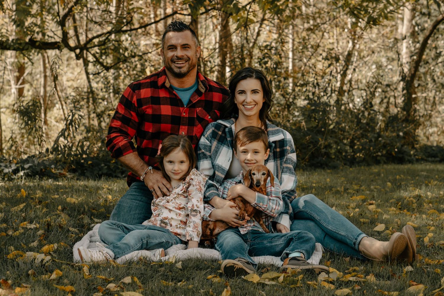 Family in coordinating plaid shirts sitting together on grass for outdoor portrait session.