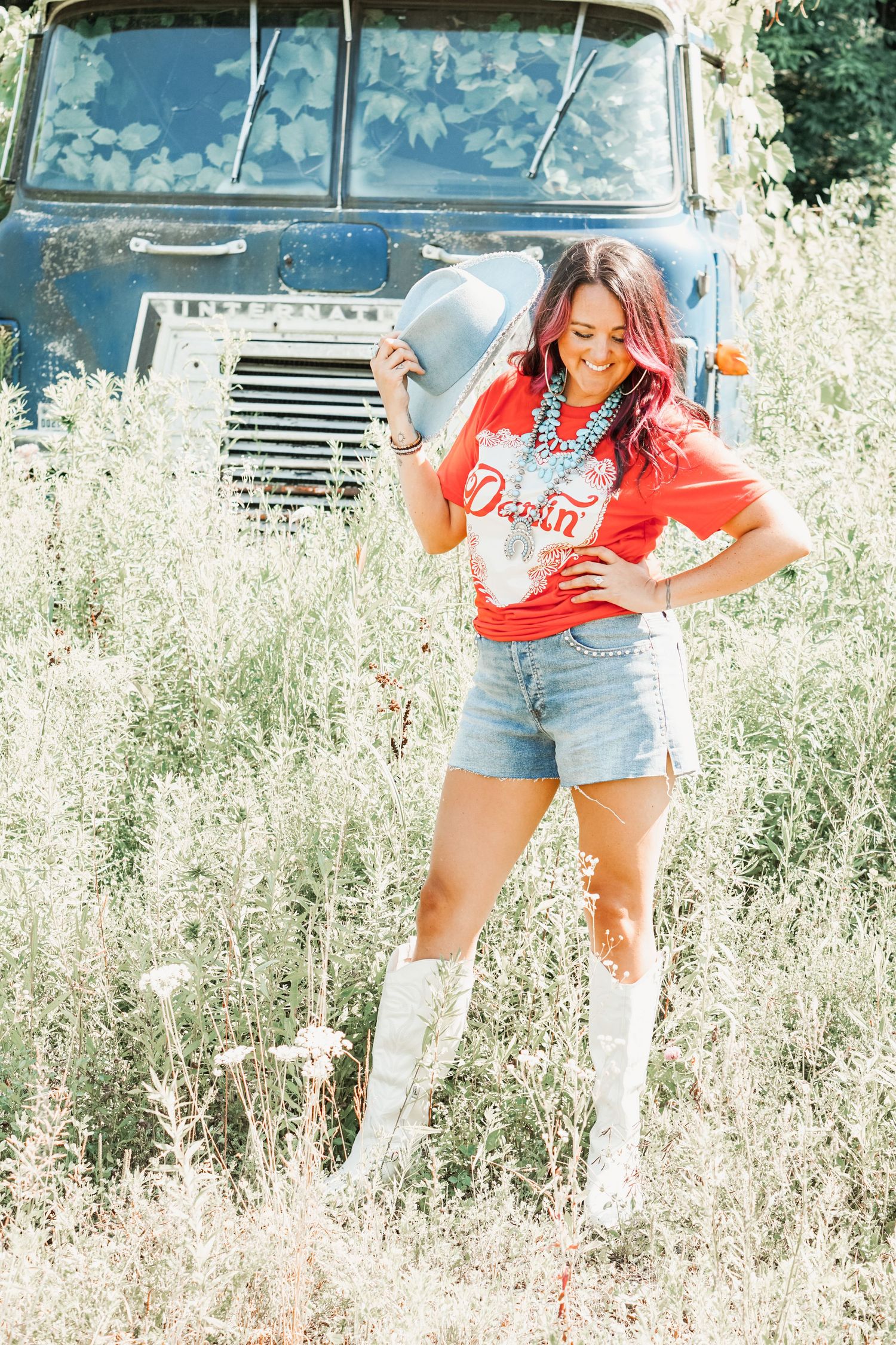 Country western fashion look with red shirt and white boots posed in front of vintage blue truck in field.