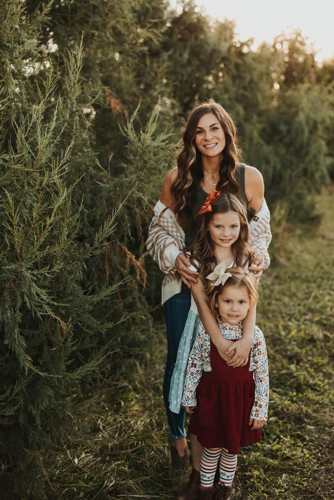 A family portrait featuring adults and children posed together among evergreen trees at sunset.