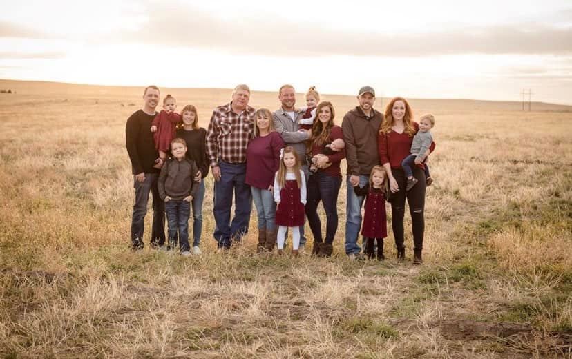 Large family group dressed in coordinating outfits poses together at sunset in open field.