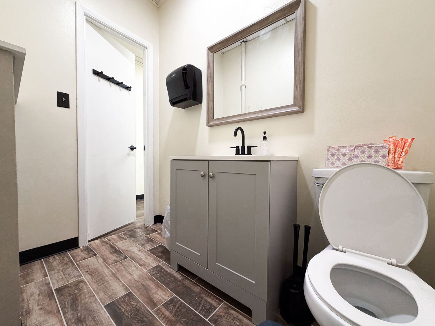 Modern bathroom with gray vanity, framed mirror, wood-look tile floors, white toilet, and door with black hardware.