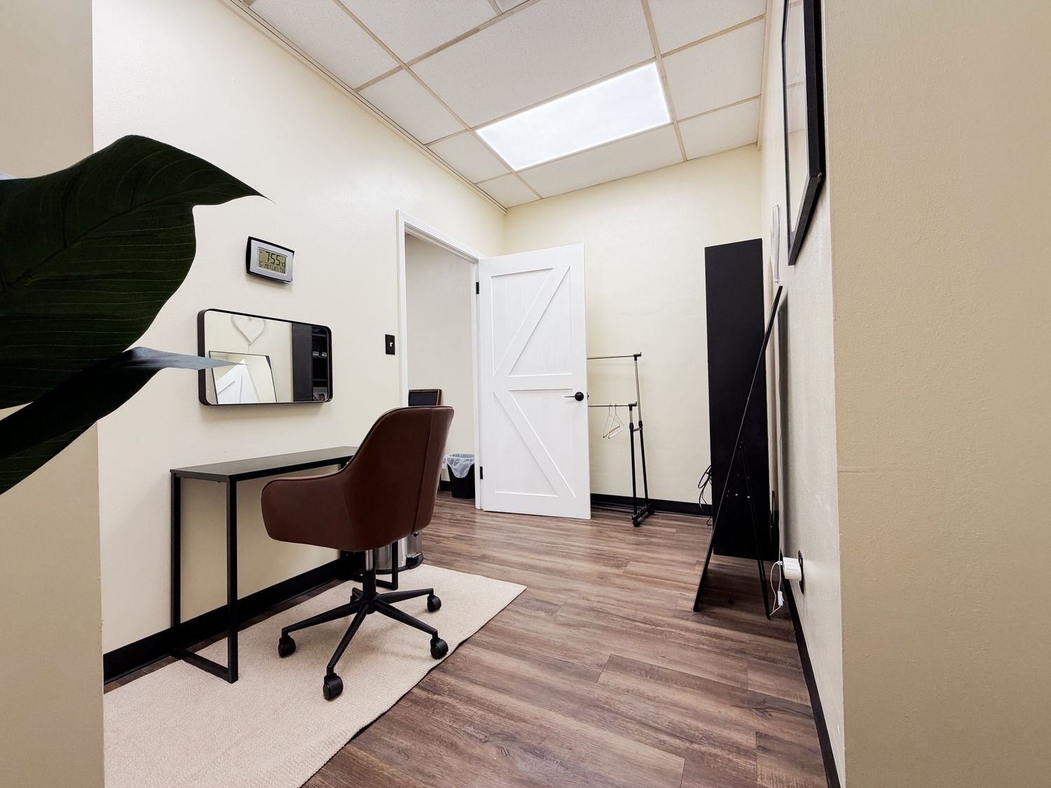 Small office room with wood floors, a brown desk chair, black desk, mirror, and white barn door.