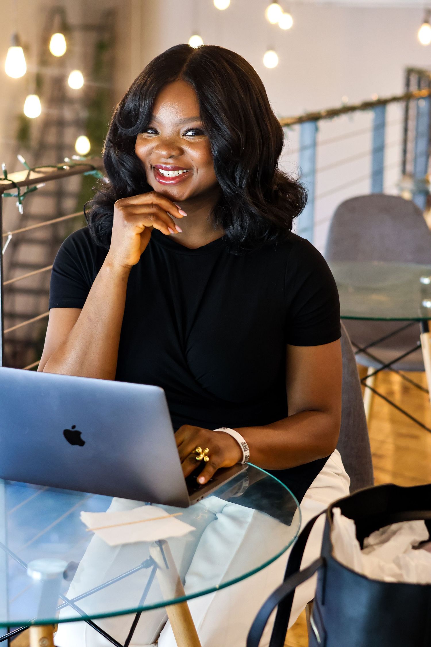 Smiling Black woman in black t-shirt working on MacBook laptop at glass desk in modern office with string lights.