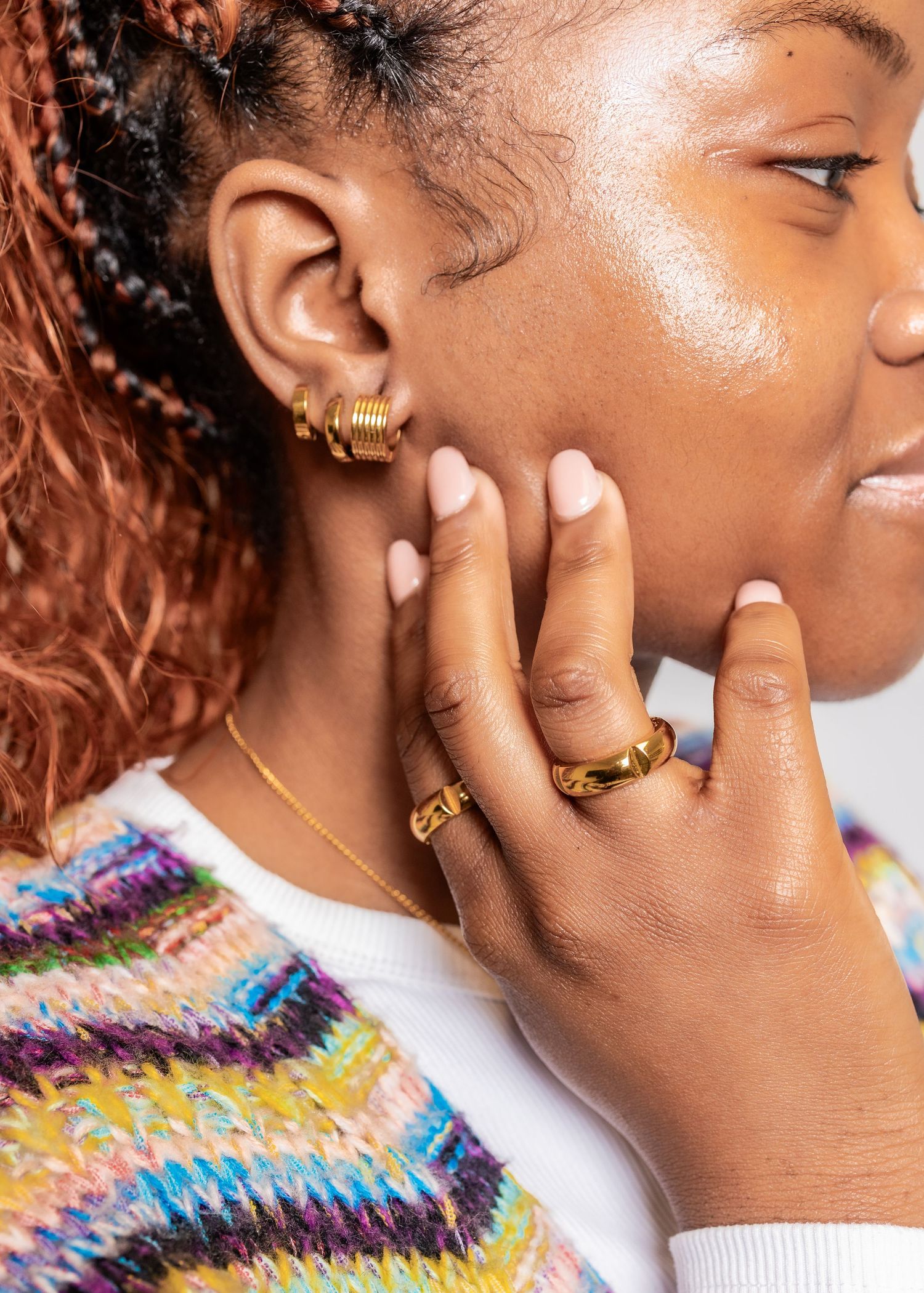 Woman wearing gold hoop earrings and a chunky gold ring, touching her face with manicured nails.