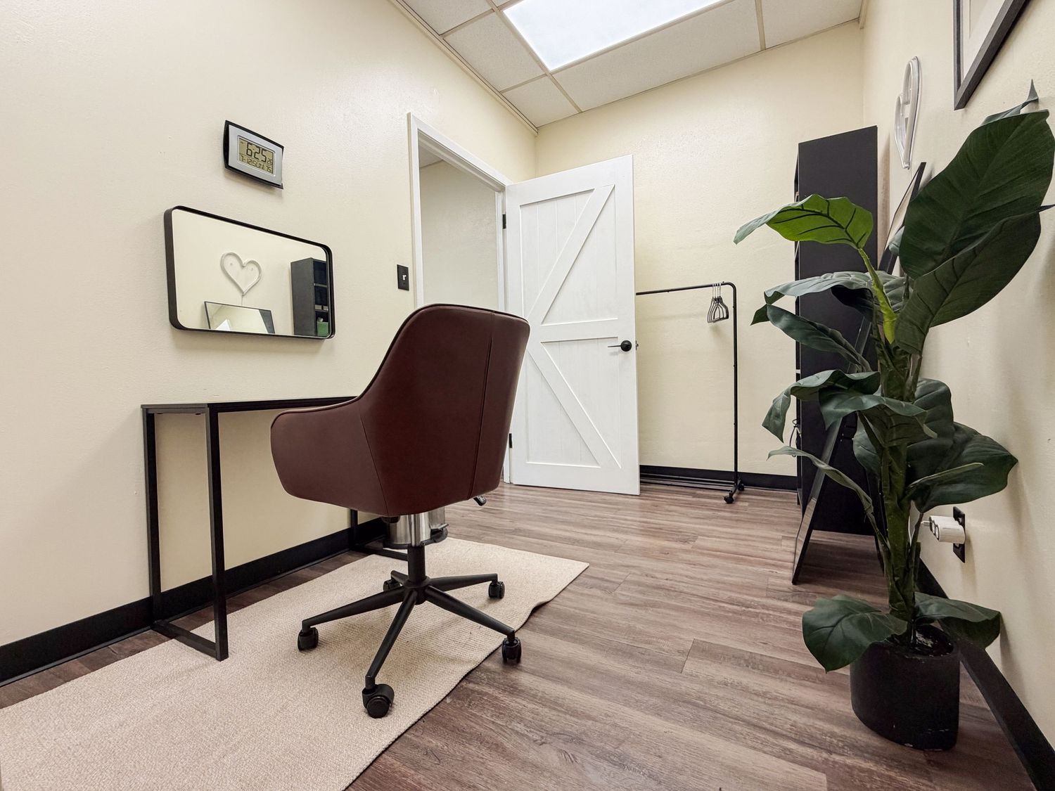 Small private office with brown leather chair, desk with mirror, hardwood floors, and decorative plant near white barn door.