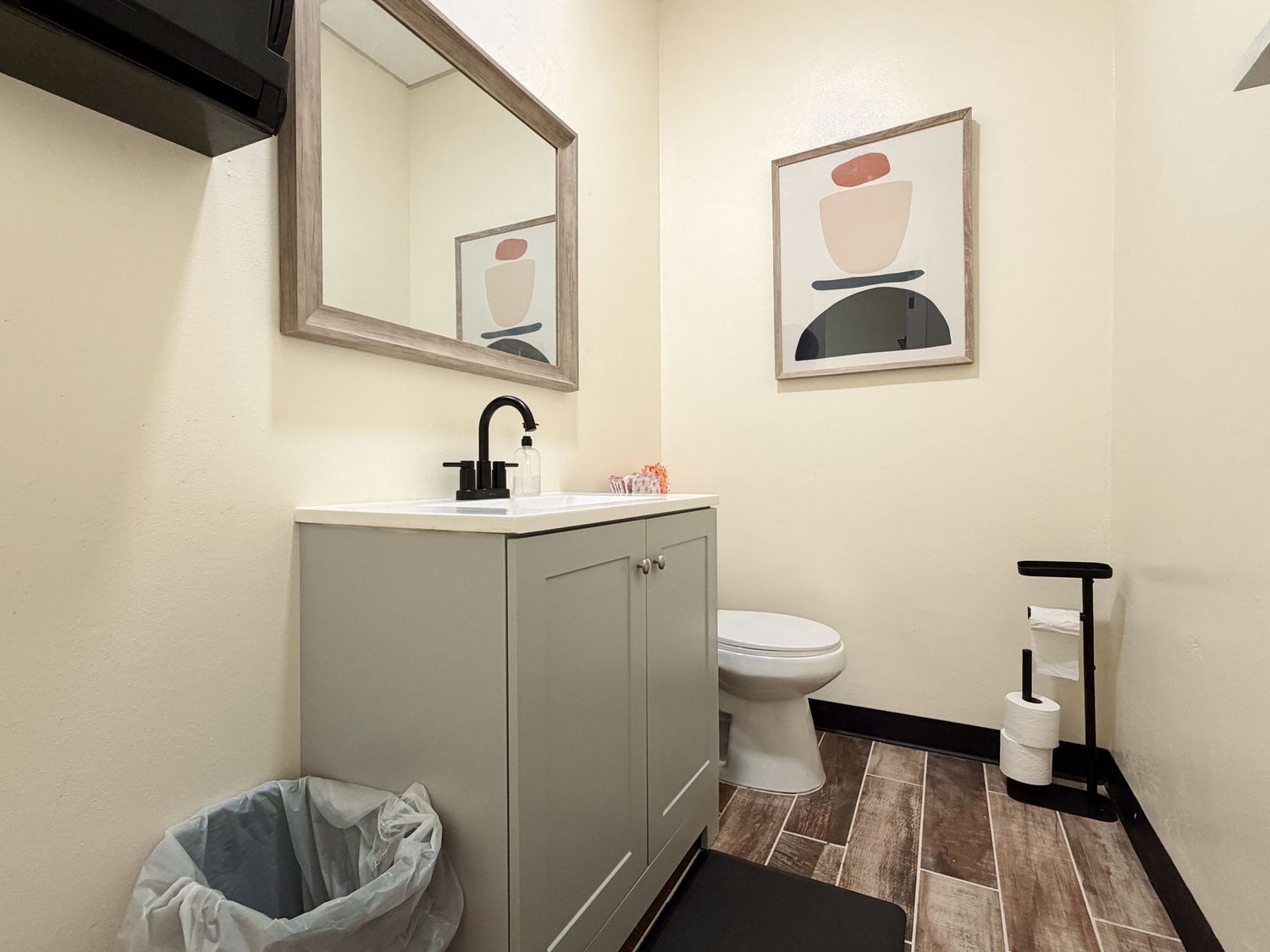 Modern bathroom with gray vanity, framed mirror, toilet, wood-look tile floors, and abstract wall art.