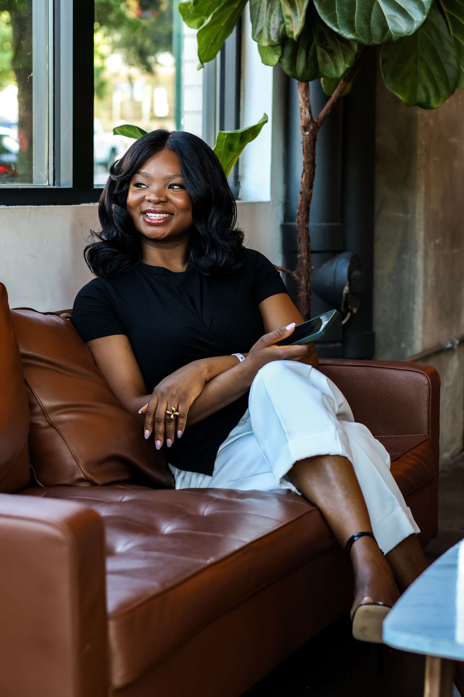 Smiling Black woman in black top and white pants sitting on brown leather sofa holding smartphone.