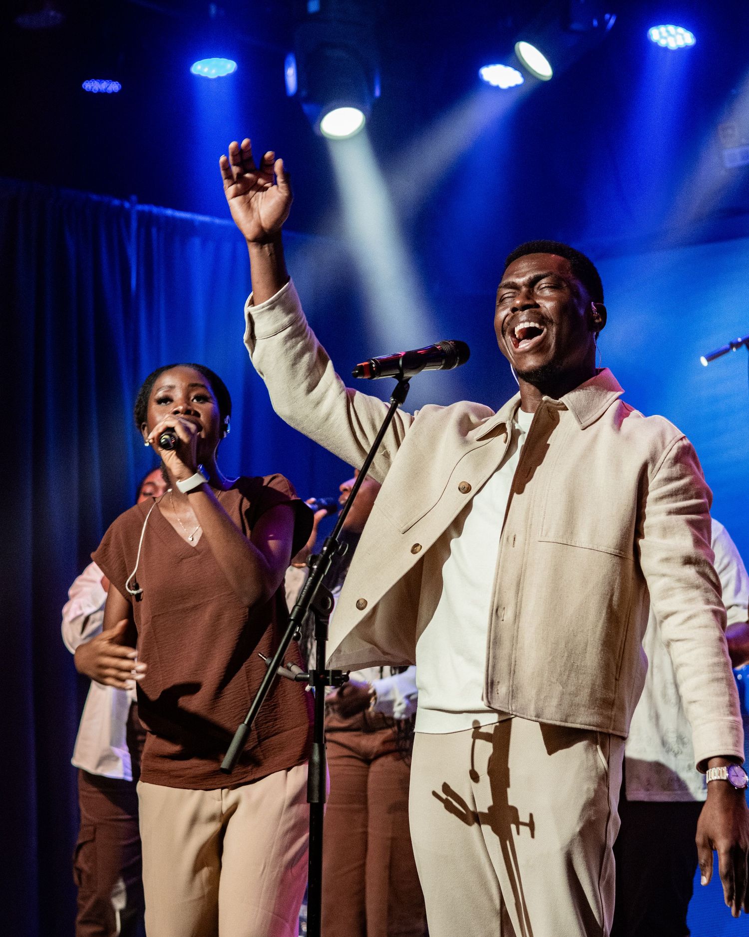 Two performers singing on stage under blue lights, one raising hand joyfully, wearing neutral-toned outfits.
