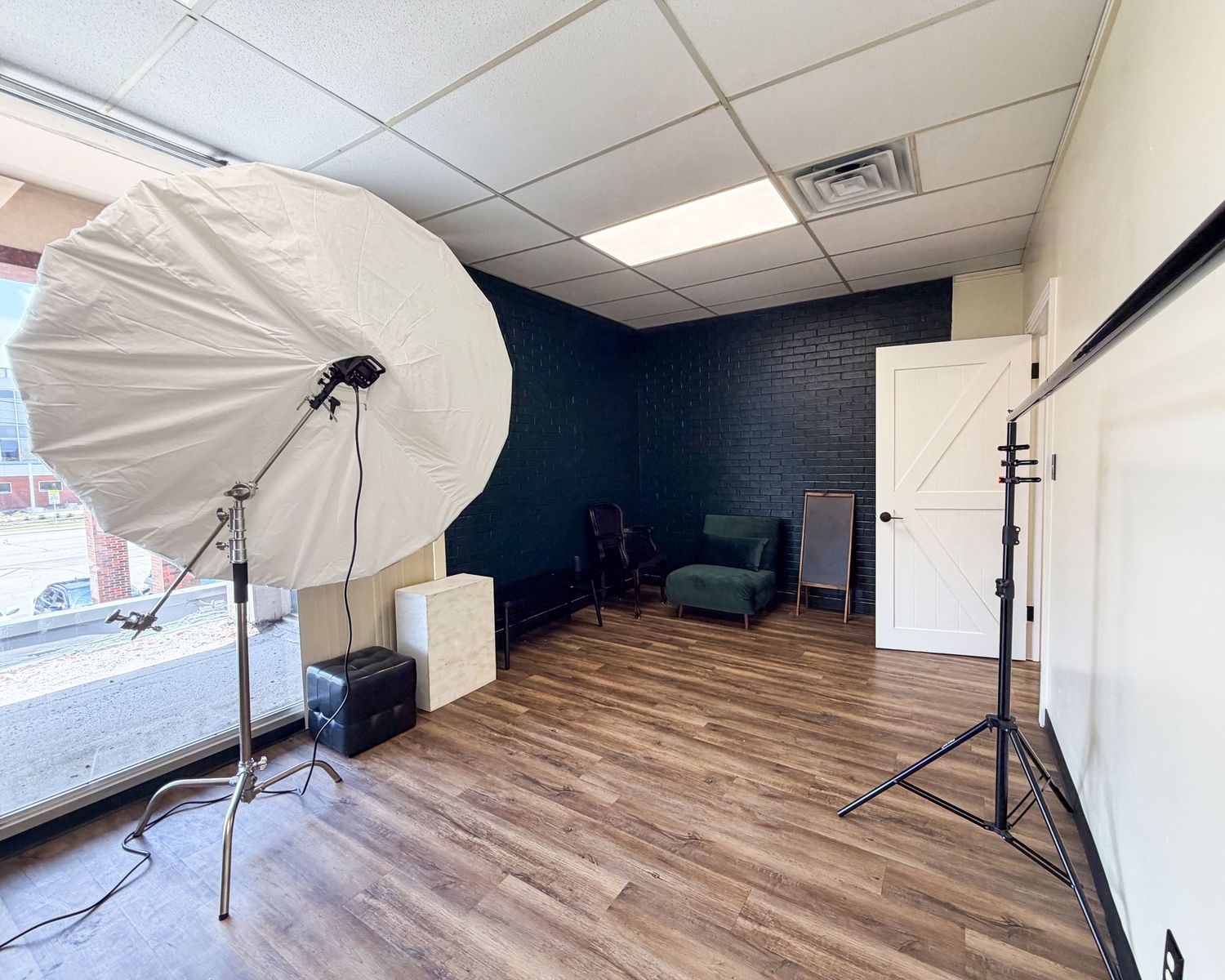 Professional photography studio with umbrella lights, hardwood floors, dark accent wall, and a cozy green chair setup.