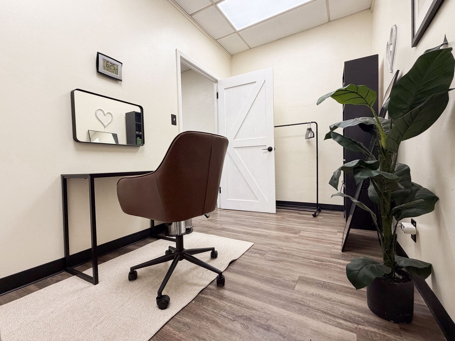 Small modern office with brown leather chair, minimalist desk, mirror, and large green plant on wood floors.