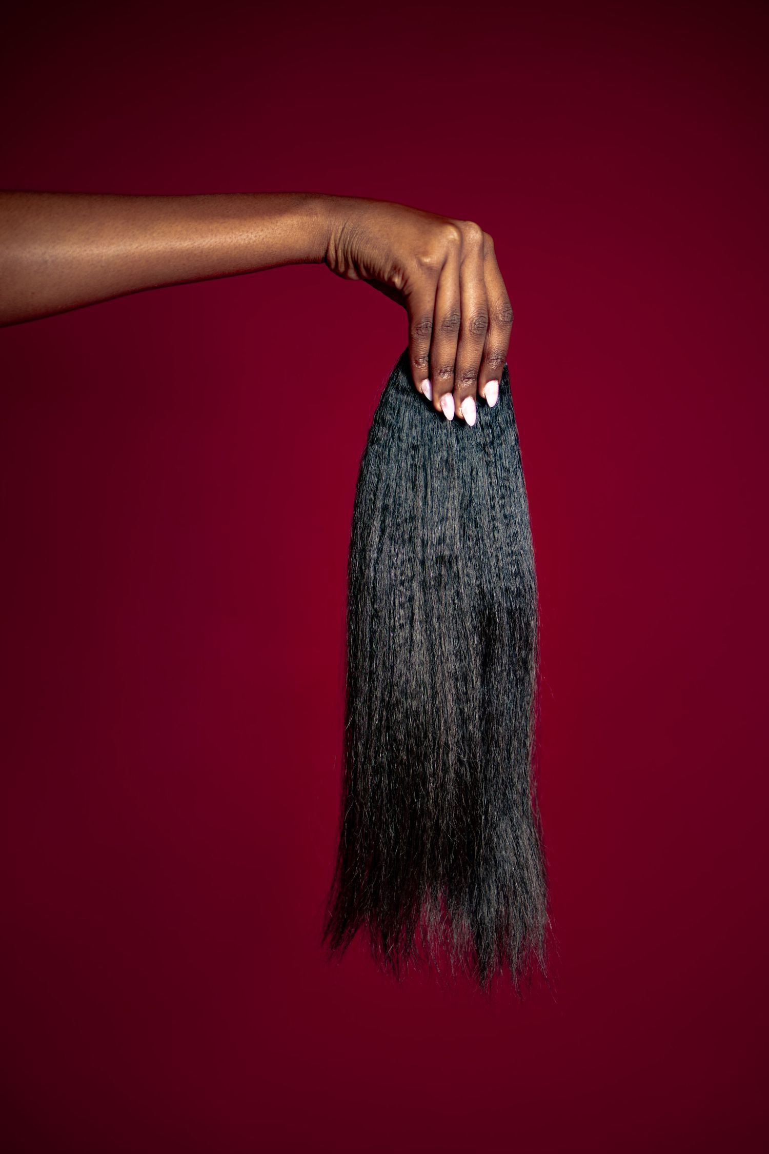 A hand with white nails holds a bundle of dark kinky straight hair extensions against a deep red background.