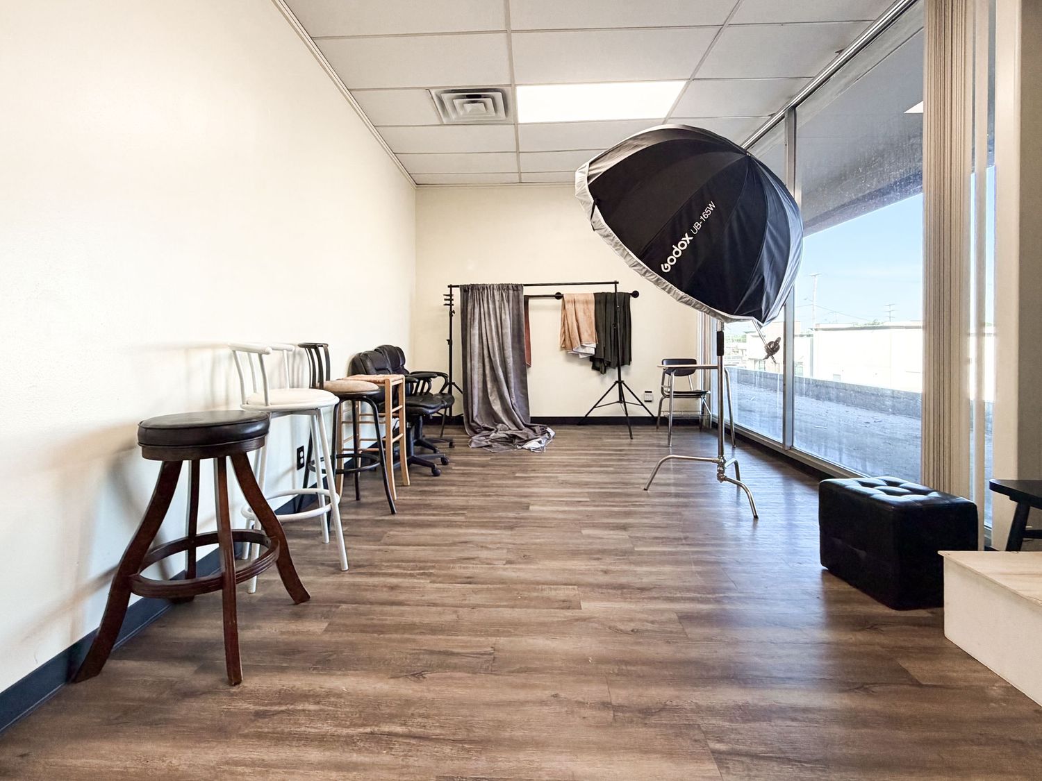 Professional photography studio with hardwood floors, umbrella light, backdrop stand, and large windows providing natural light.