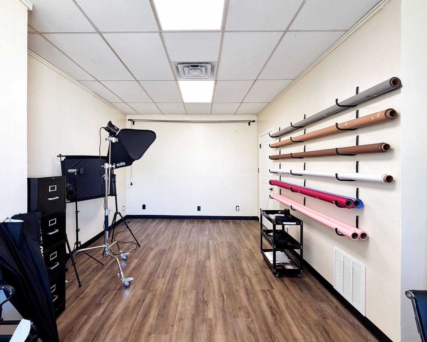 Photography studio room with lighting equipment, filing cabinet, and wall-mounted roll storage rack on hardwood floors.