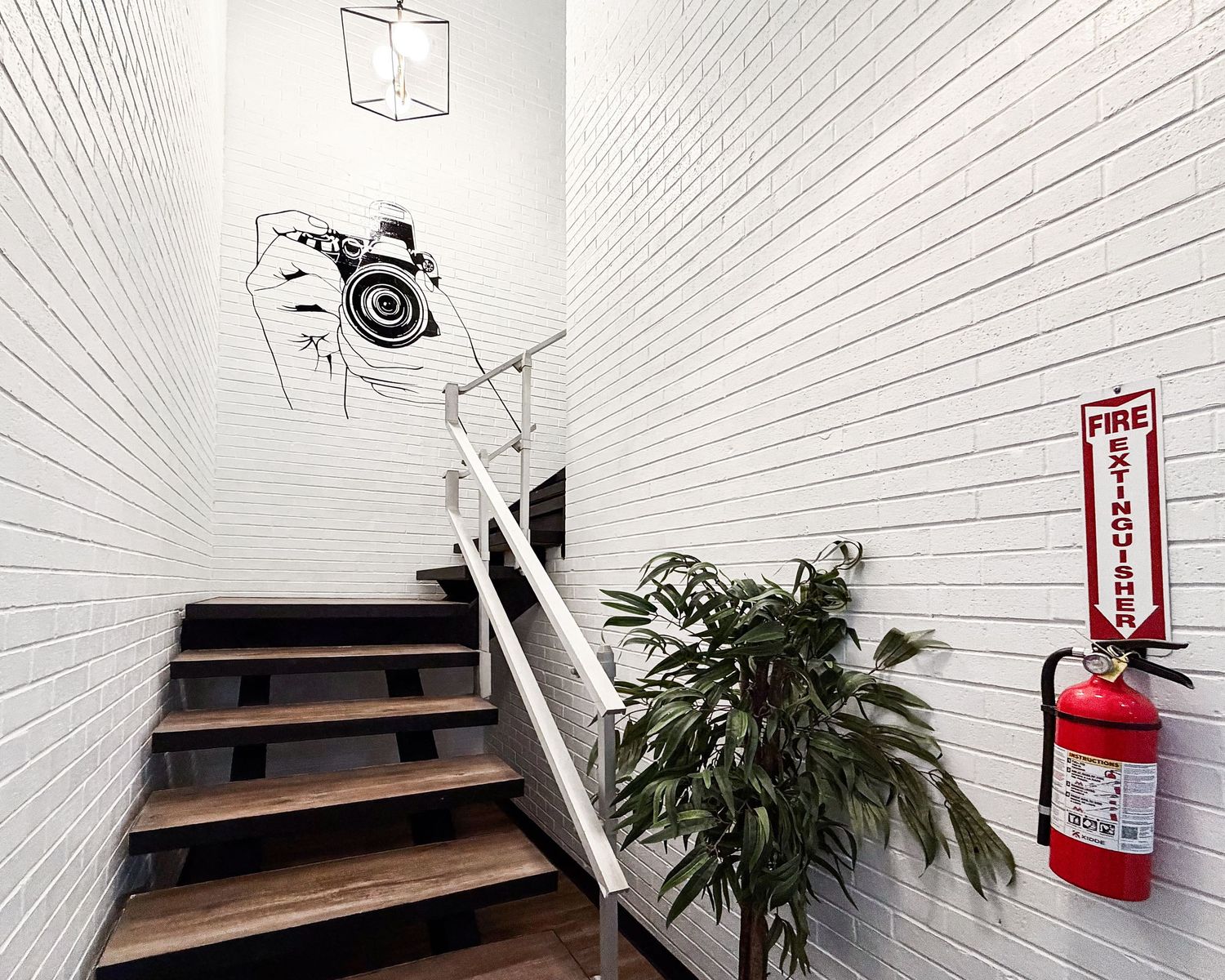 Modern staircase with white brick walls, wood steps, camera wall art, green plant, and red fire extinguisher.