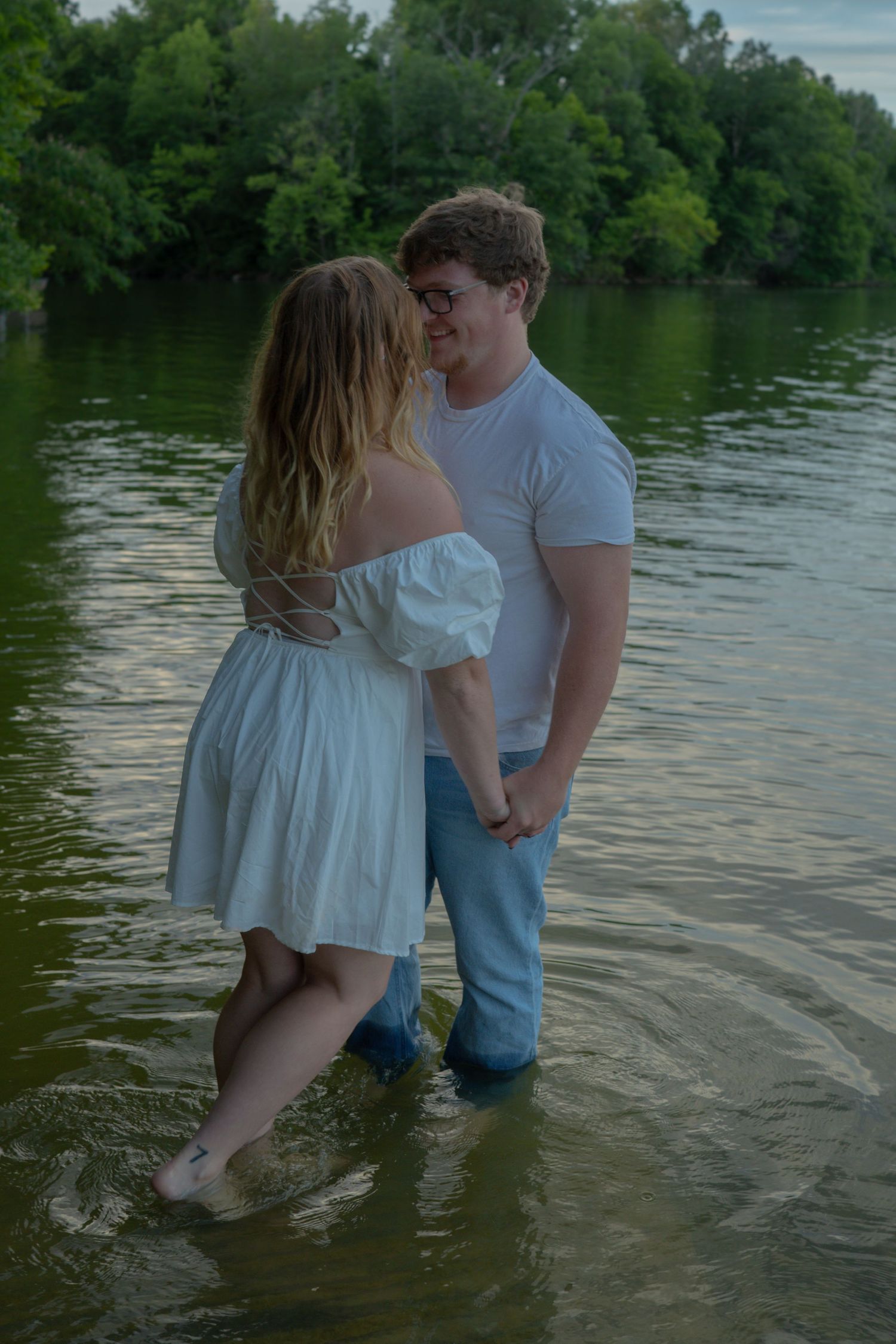 A romantic couple stands together in shallow lake water, with lush green trees reflecting on the calm surface at sunset.