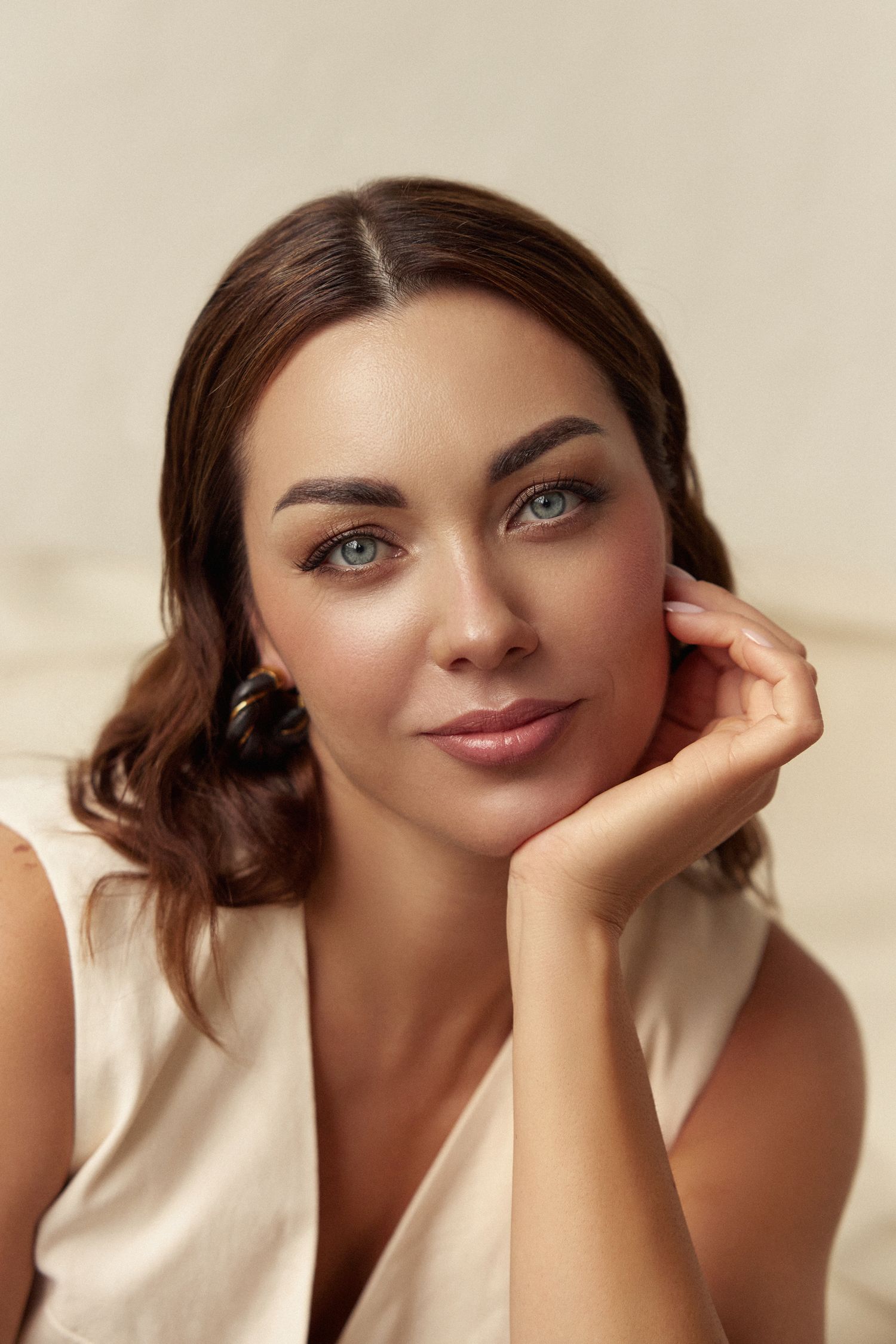 Elegant brunette woman with blue eyes resting chin on hand, wearing a cream sleeveless top against a neutral background.