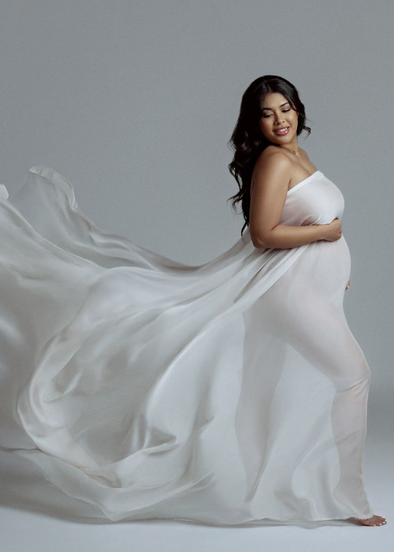 Beautiful pregnant woman smiles in flowing white chiffon maternity gown with fabric trailing in studio.