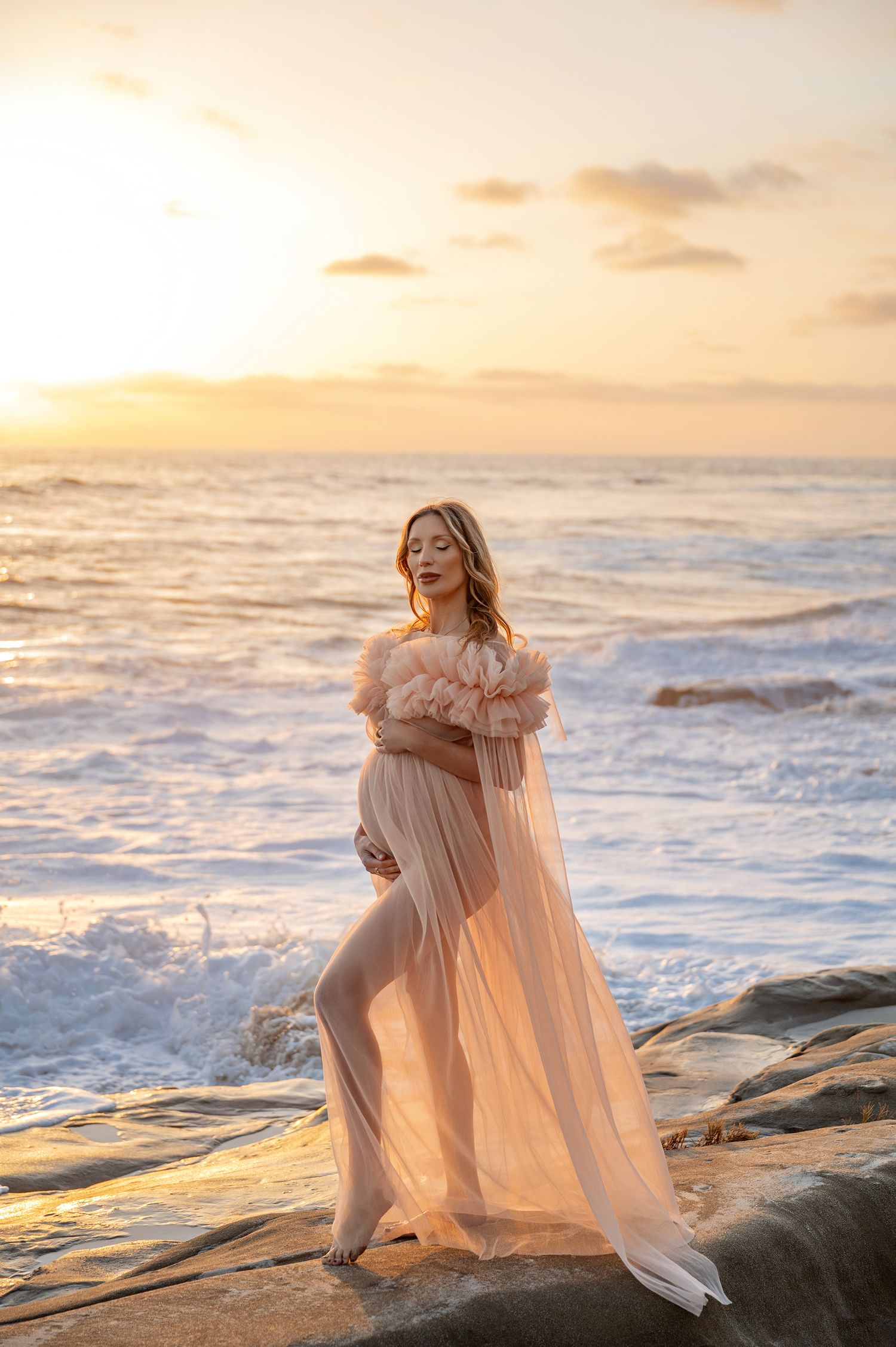 A pregnant woman in a sheer peach off-shoulder gown stands on ocean rocks at sunset.
