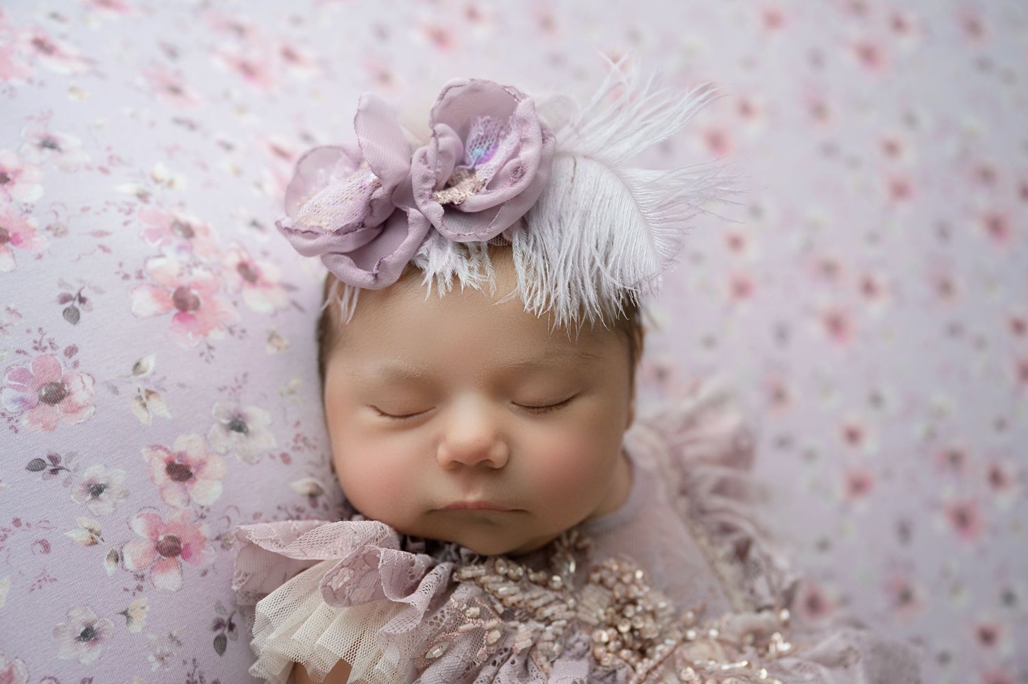 Sleeping newborn baby wearing a lavender floral headband on a pink floral backdrop.
