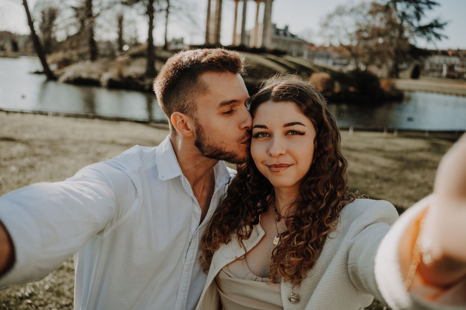 Couple taking a romantic selfie outdoors by a lake, man kissing woman on cheek, sunny day in a park.