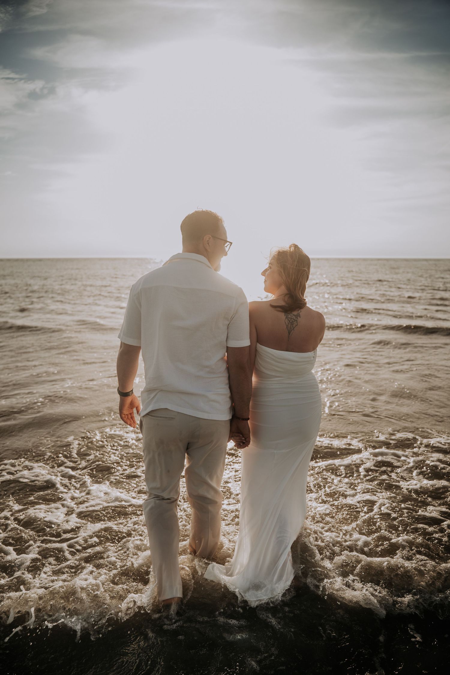 Couple in white outfits holding hands standing in ocean waves at sunset, romantic beach photo session.