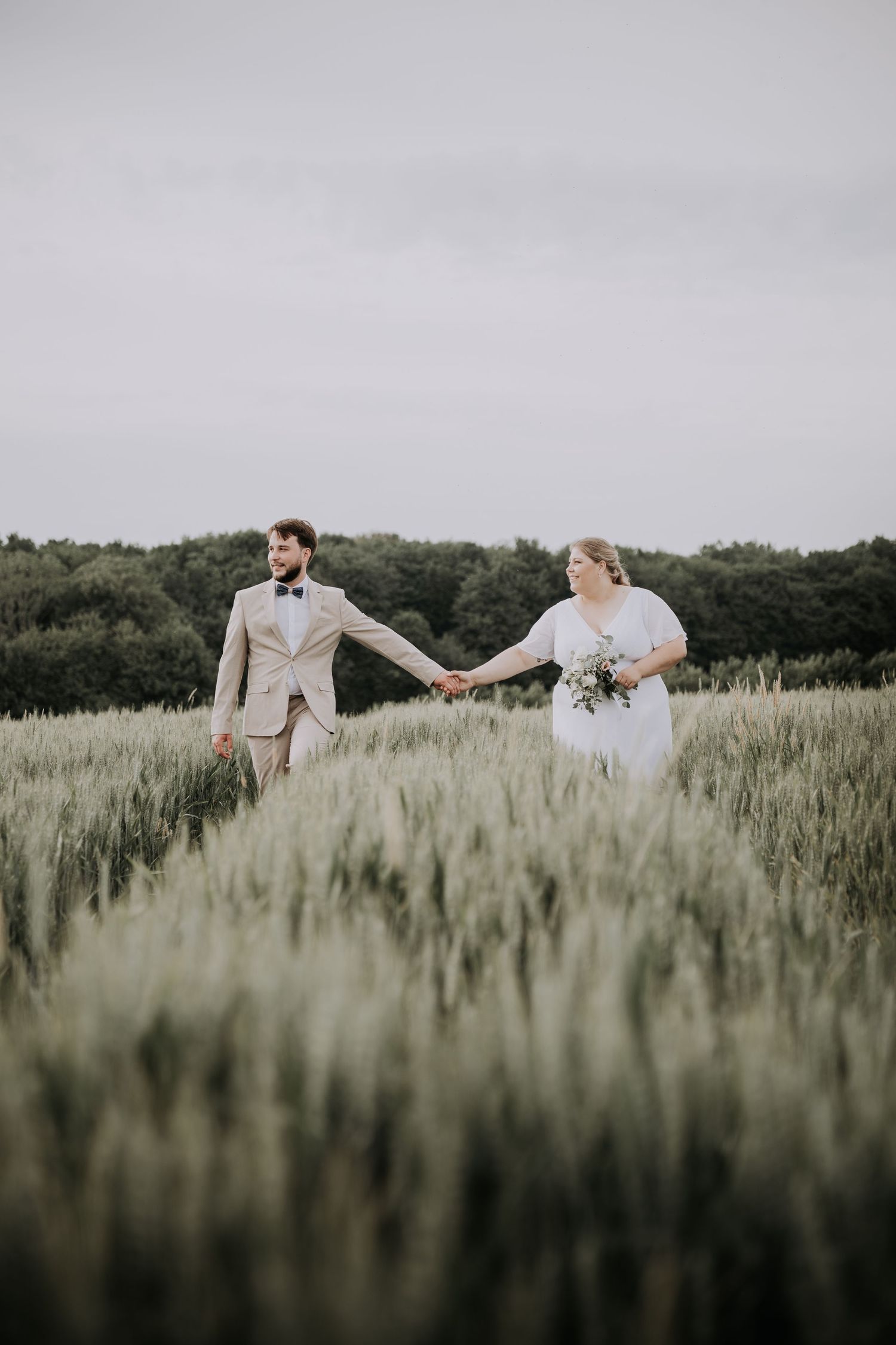 Bride and groom holding hands in a lush green wheat field, groom in beige suit, bride in white dress holding bouquet.