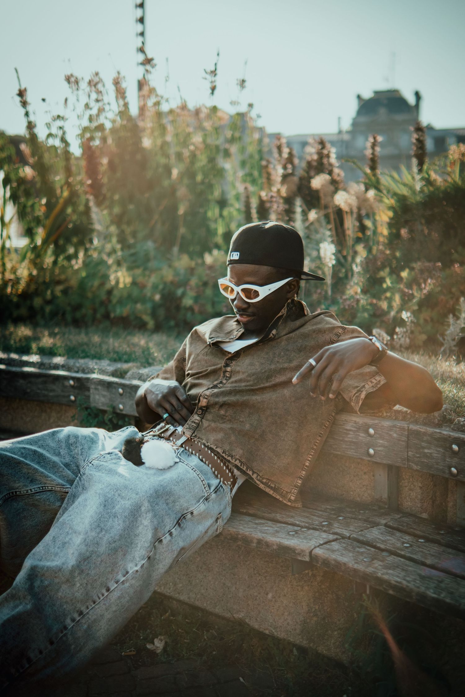 Man in black cap and white sunglasses relaxing on a bench surrounded by lush greenery in warm sunlight.