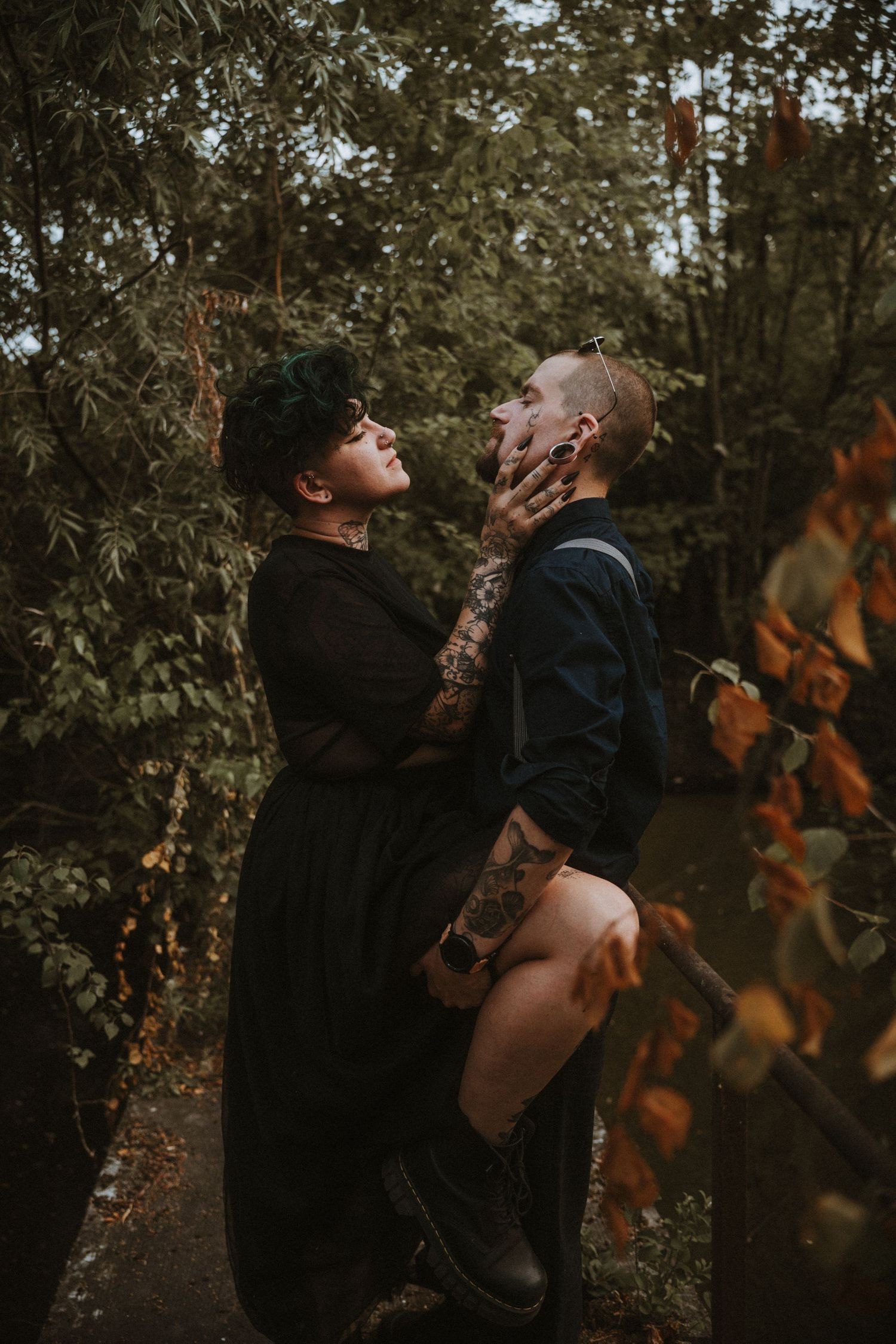 Tattooed couple sharing romantic moment in autumn forest, woman in black dress holding partner's face tenderly.