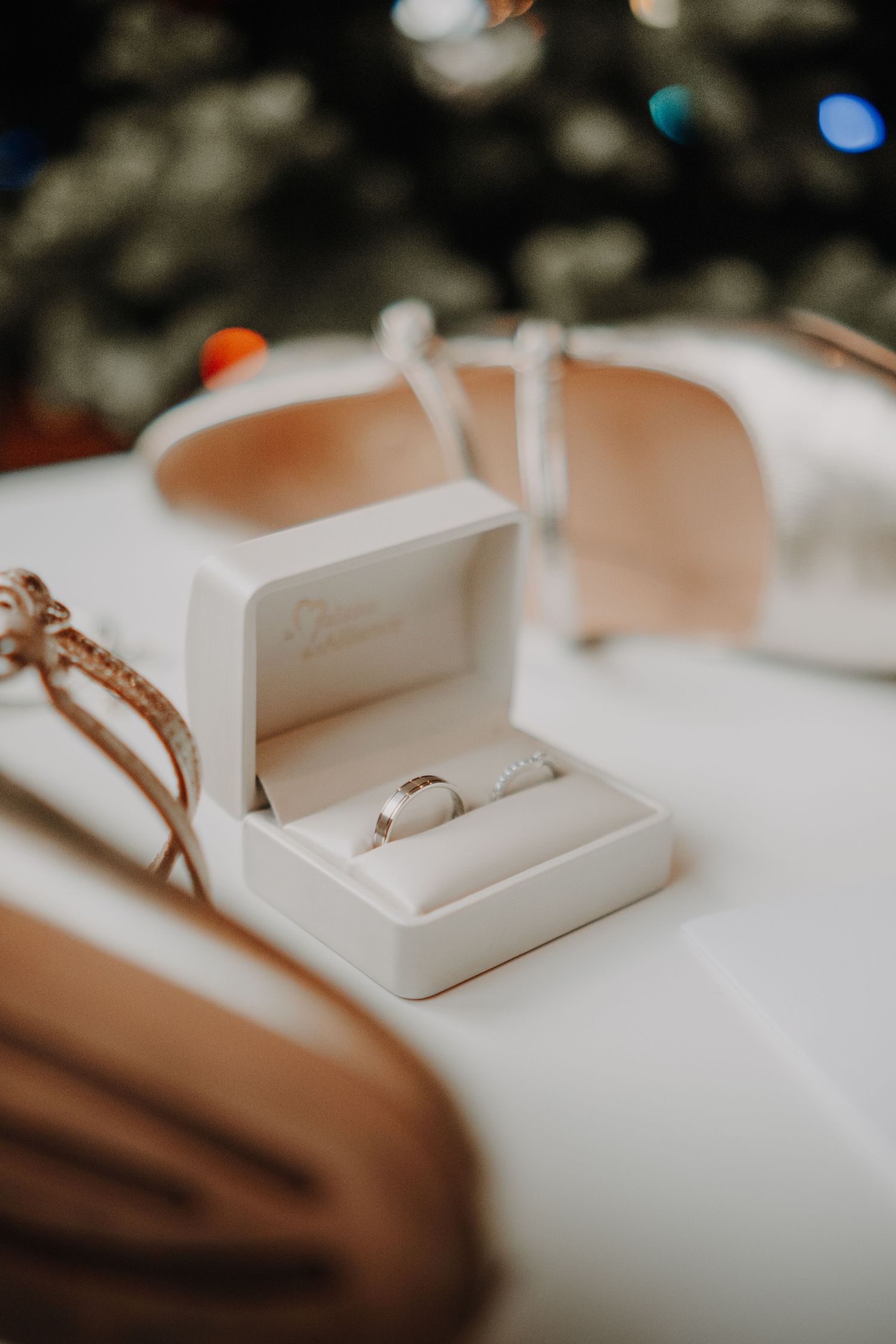 Silver diamond engagement ring displayed in a white velvet jewelry box on a white surface with bokeh lights in background.