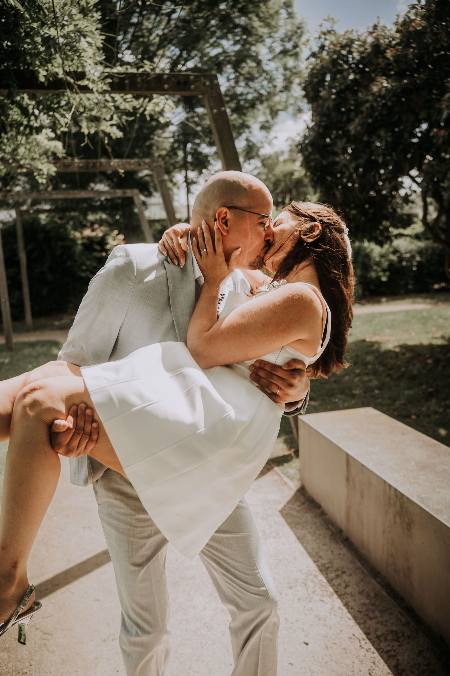 A groom in a light suit lifts his bride in a white dress as they share a romantic kiss outdoors.