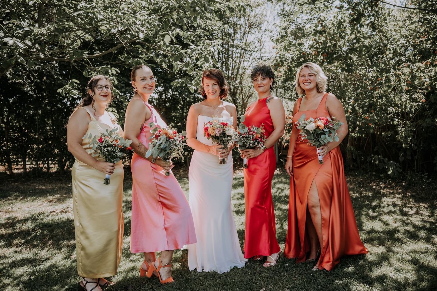Bride in white gown poses with five bridesmaids in colorful satin dresses holding bouquets outdoors.