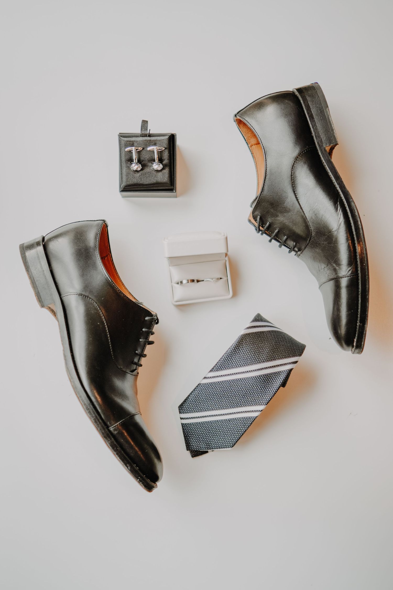 Black leather dress shoes, striped tie, cufflinks, and wedding rings arranged on white background for groom wedding accessories.